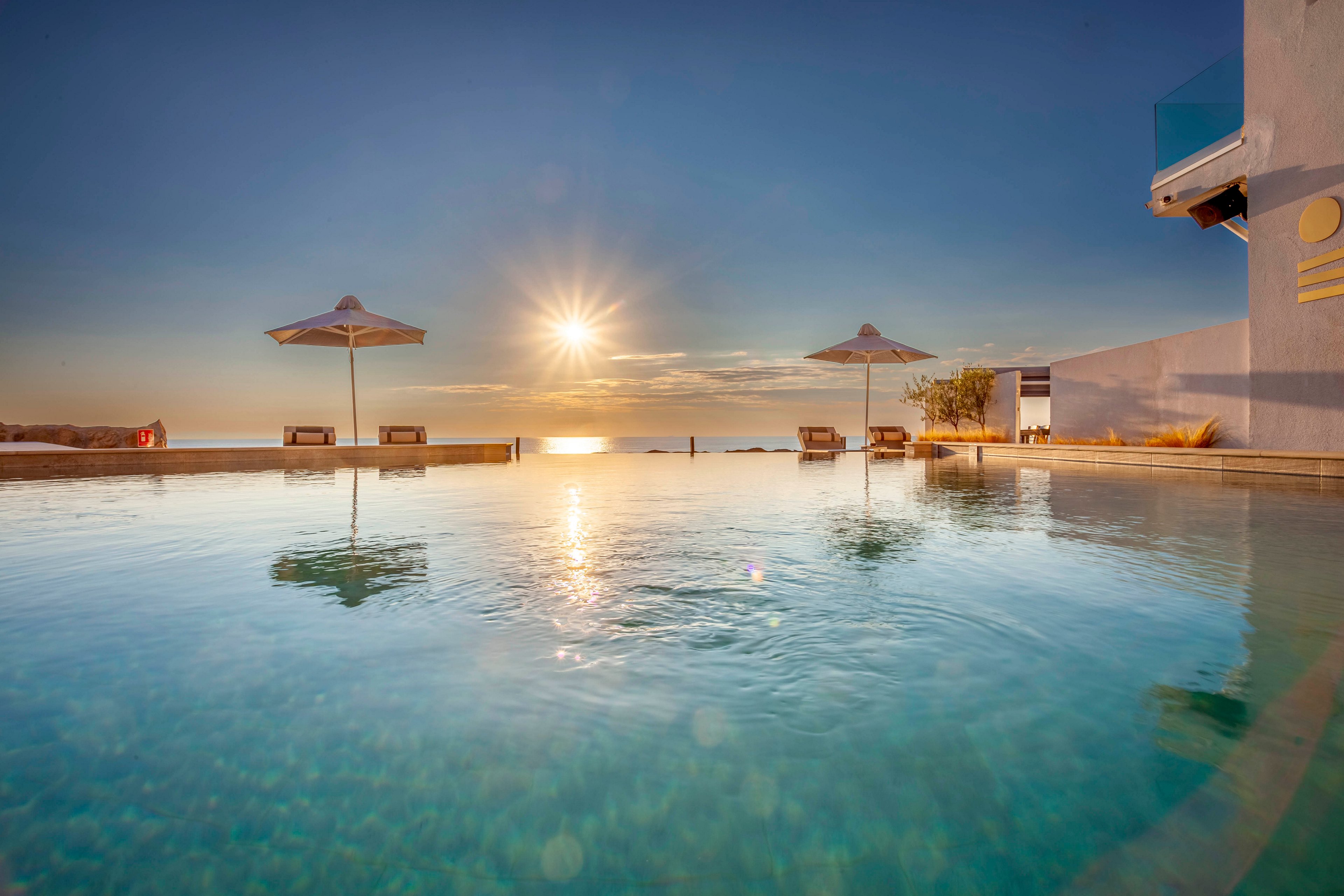 A serene infinity pool overlooks the ocean as the sun sets, casting warm golden light across the water. Two white umbrellas and several lounge chairs are positioned along the pool's edge, creating a tranquil atmosphere. The scene features clear skies and a modern architectural backdrop, emphasizing relaxation and luxury. No visible text or numeric values are present in the image.