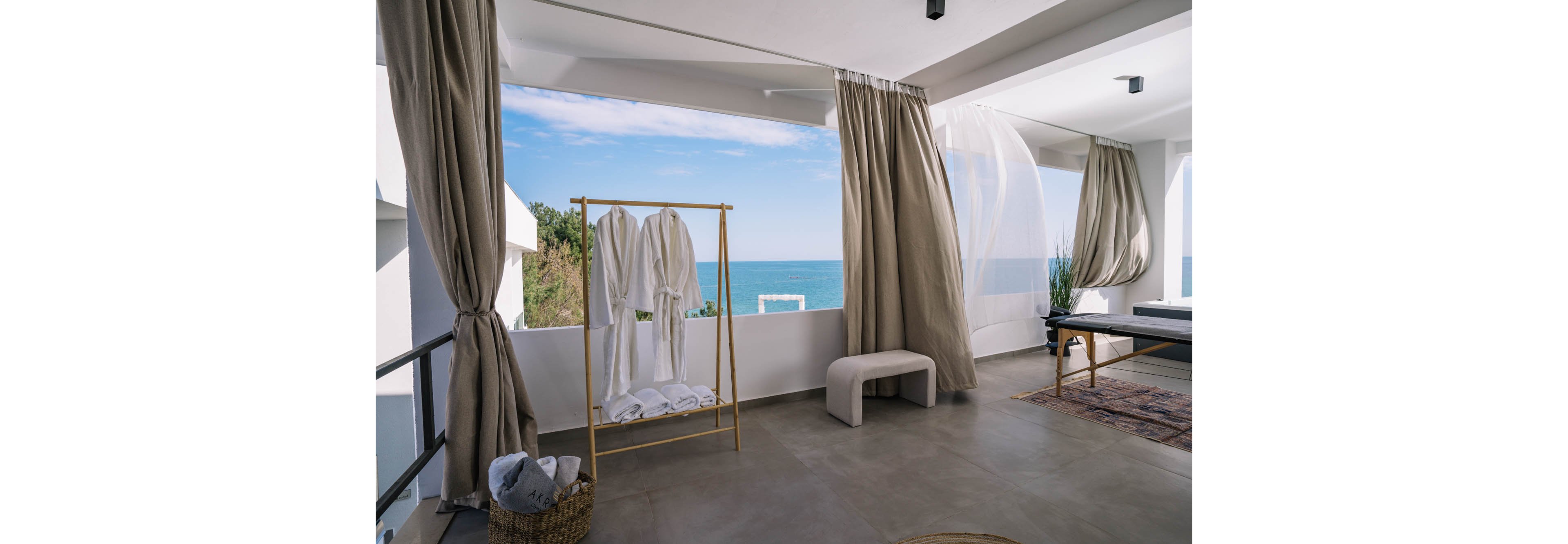 A contemporary hotel suite features a rack with white bathrobes and towels, set against large windows overlooking the ocean. Neutral curtains frame the view, and a minimalist bench sits nearby. The space is bright, airy, and decorated in soft beige and white tones, evoking a tranquil, luxurious mood.