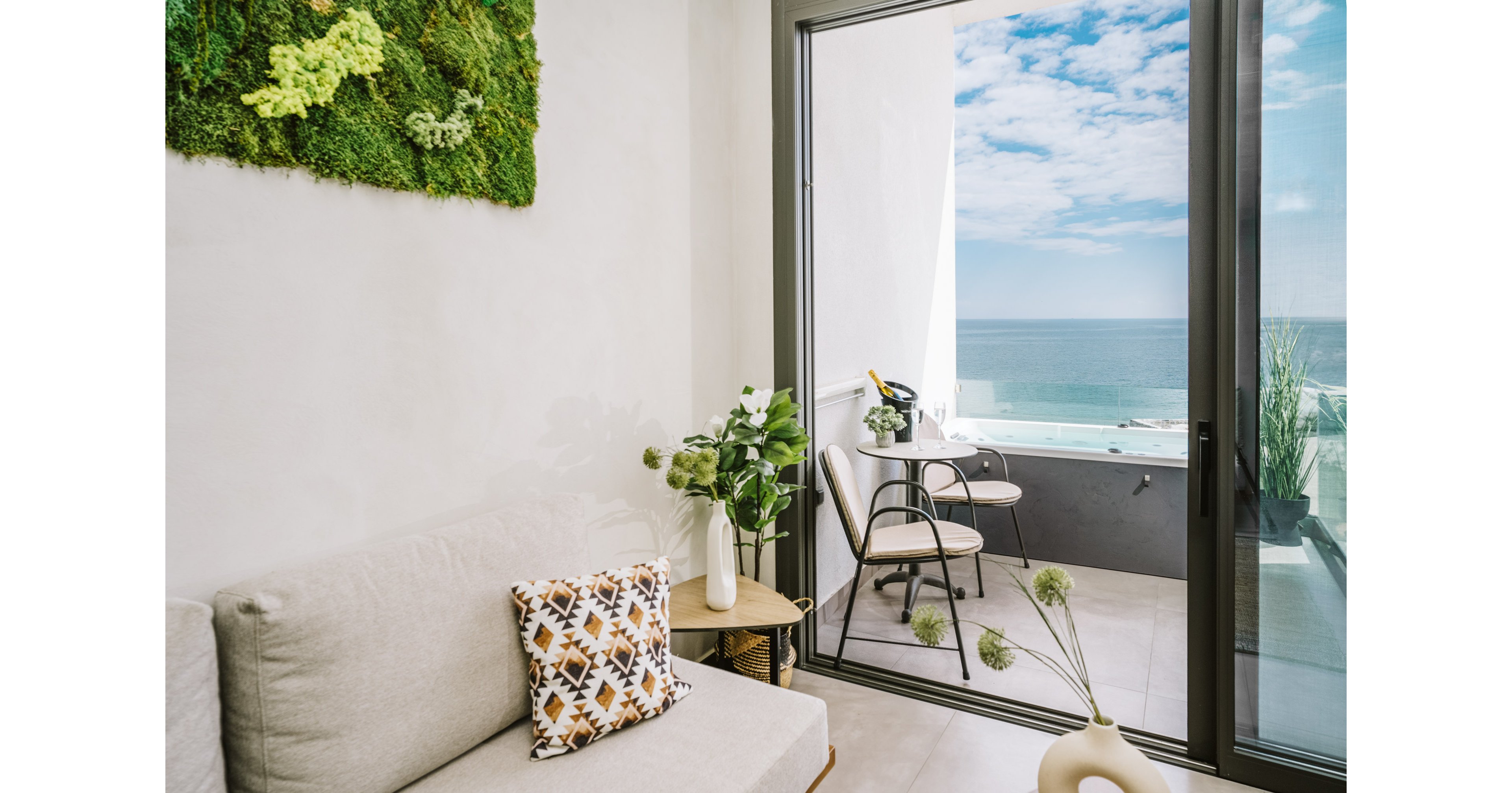 A contemporary living room opens onto a balcony overlooking the ocean, featuring a glass sliding door. The interior includes a light sofa, patterned pillow, and a vase with greenery, while the balcony has minimalist chairs and a small table. Natural light fills the space, highlighting the green wall art and serene seaside atmosphere.