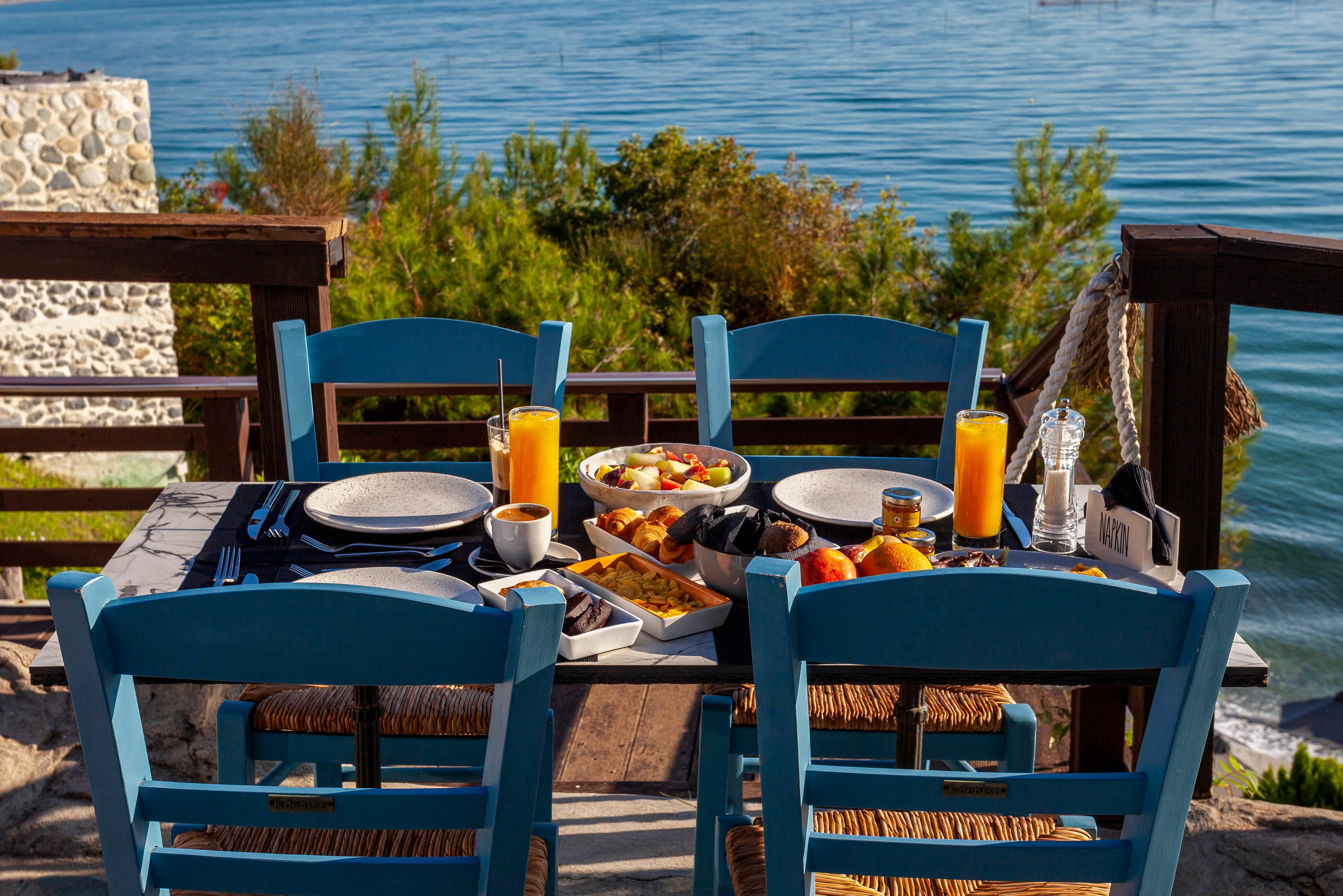 A wooden table set for breakfast is placed outdoors overlooking a calm sea. The table features blue chairs, plates, glasses of orange juice, and a variety of pastries and fruits. The setting is bright and inviting, with natural sunlight and a scenic coastal backdrop. No visible text or numbers are present in the image.