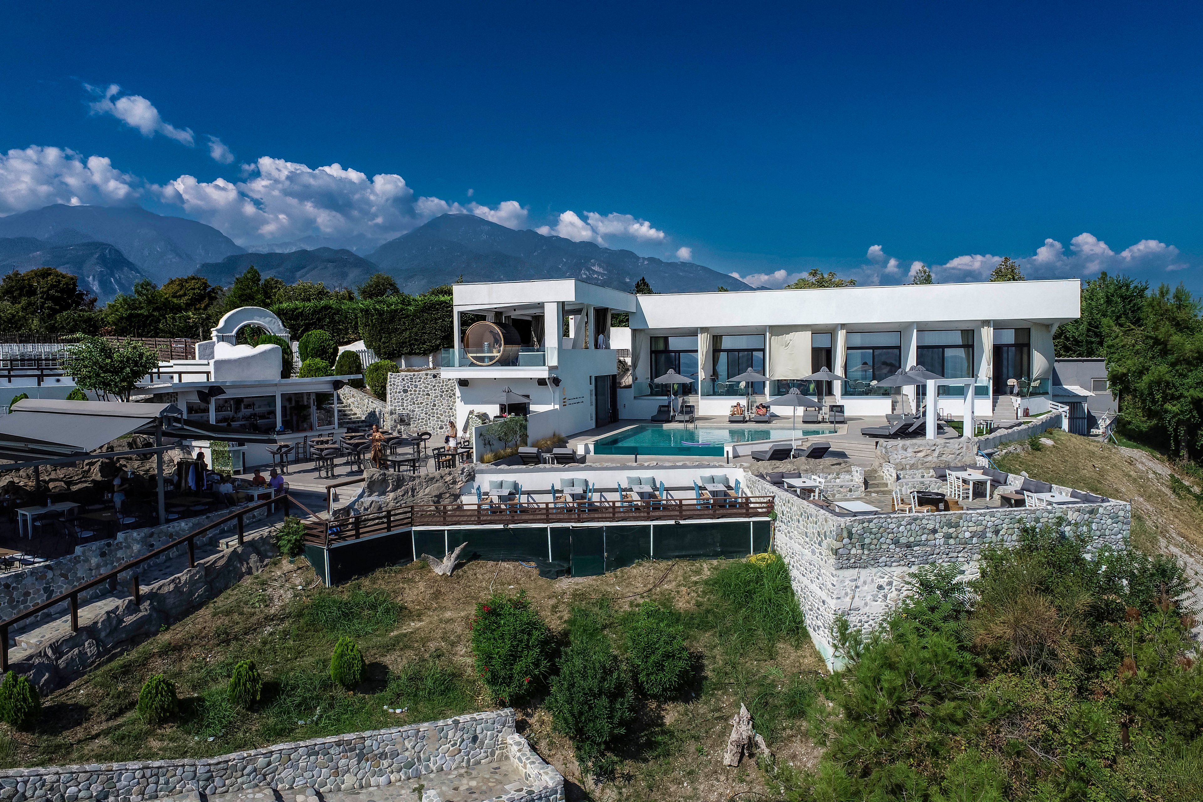 A contemporary villa features sleek white architecture and expansive glass windows, set against a backdrop of lush greenery and distant mountains. The property includes a large outdoor swimming pool, sun loungers, and multiple terraces, creating a relaxing and upscale atmosphere. The clear blue sky and landscaped grounds enhance the sense of tranquility and exclusivity.