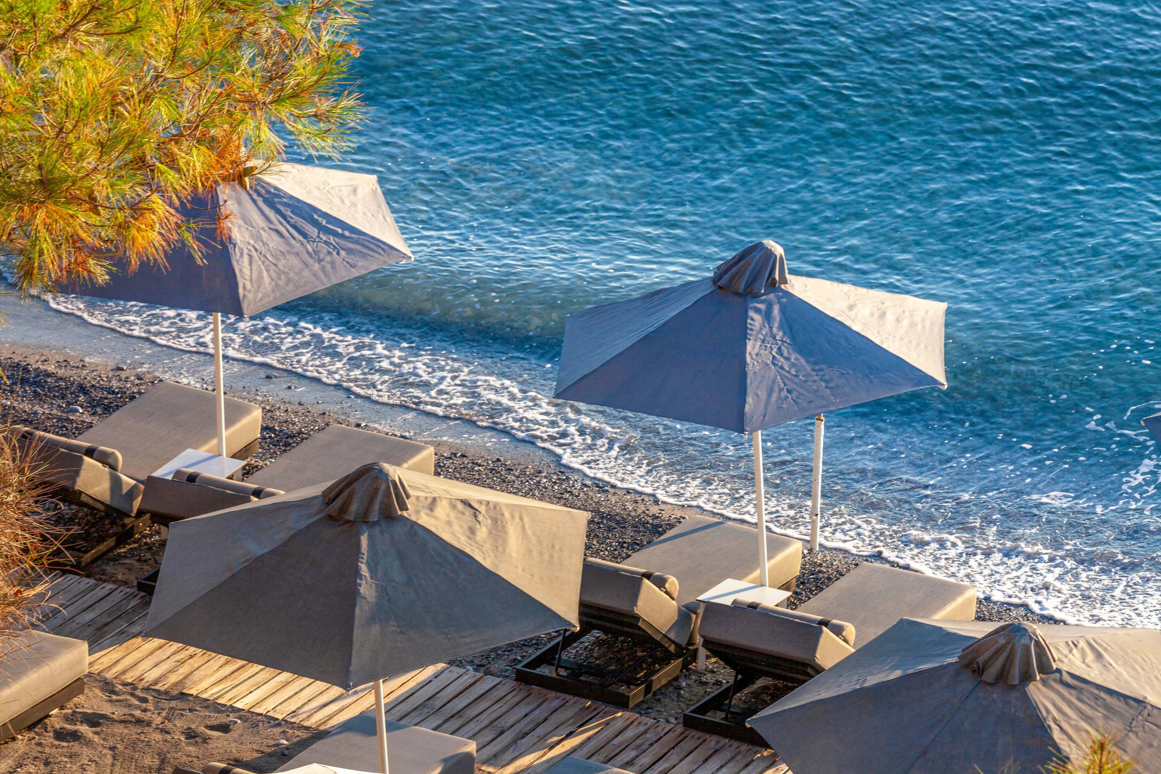 Several gray sun umbrellas and loungers are arranged on a wooden deck overlooking a calm shoreline. The scene is peaceful, with gentle waves meeting the dark sand and clear blue water. No people are present, and the setting is shaded by a nearby tree, evoking a relaxed, serene mood.