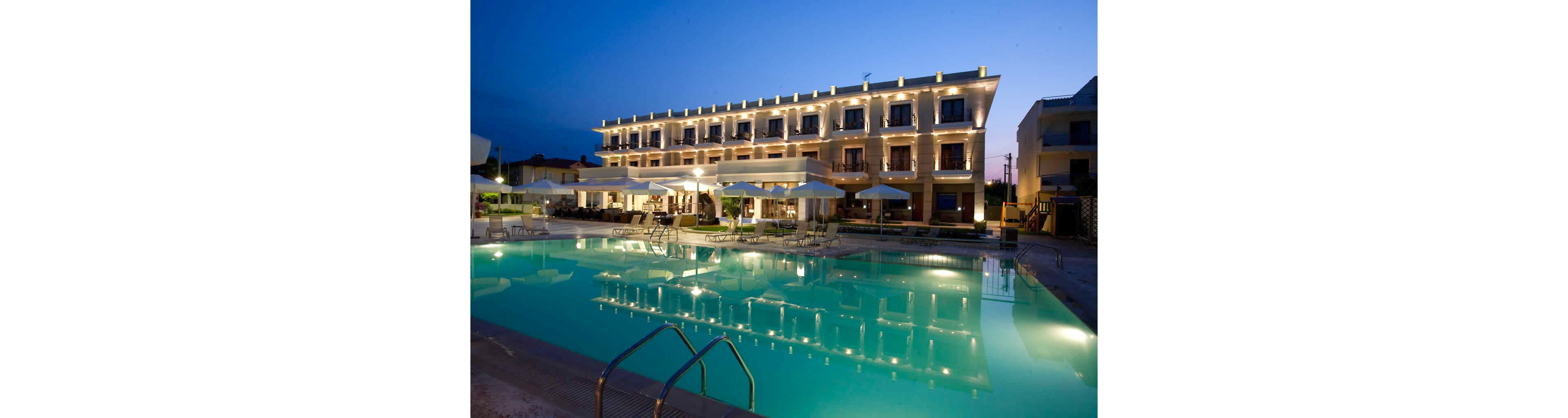 A contemporary hotel building is shown at twilight, with warm exterior lighting highlighting its architecture. The foreground features a large swimming pool reflecting the lights and structure, surrounded by poolside umbrellas and lounge chairs. The scene evokes a tranquil, inviting atmosphere suitable for leisure and relaxation, with a clear blue sky transitioning to night.