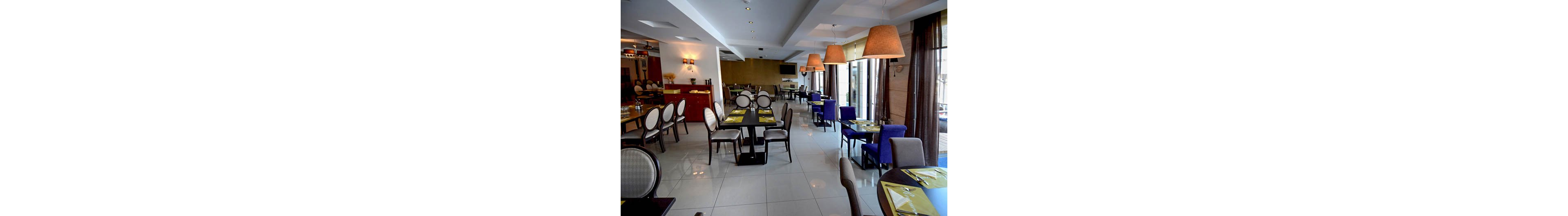 A contemporary restaurant dining area features neatly arranged tables and chairs with a mix of round and square backs. The setting is well-lit with large pendant lamps and natural light streaming through floor-to-ceiling windows. The color palette includes neutral tones with accents of blue and green from the placemats. The space appears clean, inviting, and ready for guests, with no people currently present.