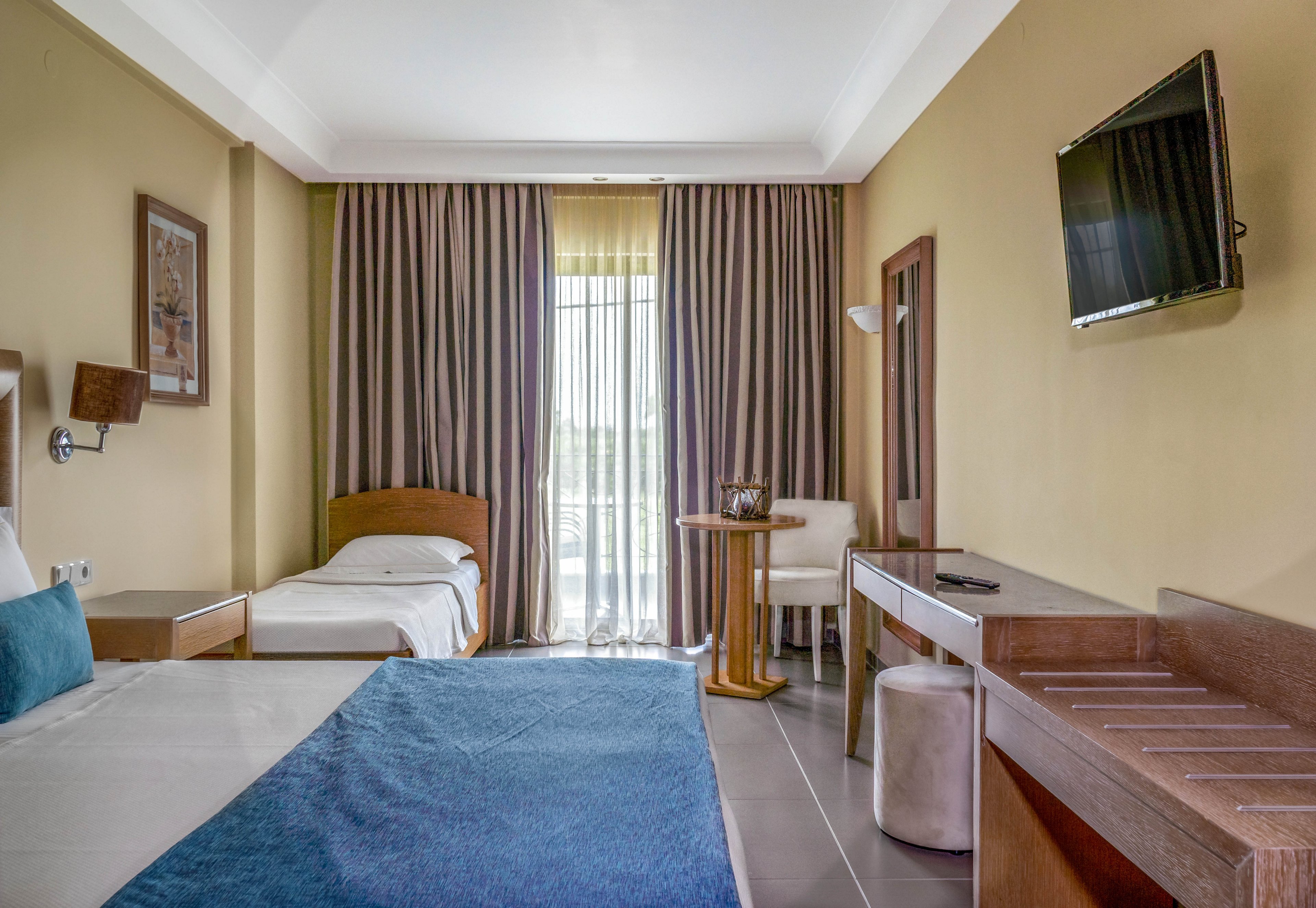 A contemporary hotel room features a double bed and a single bed, both with blue accents. The space includes a wall-mounted television, a desk, and a small seating area near a window with sheer curtains. Neutral tones and natural light create a calm, inviting atmosphere. No visible text or numeric values are present in the image.