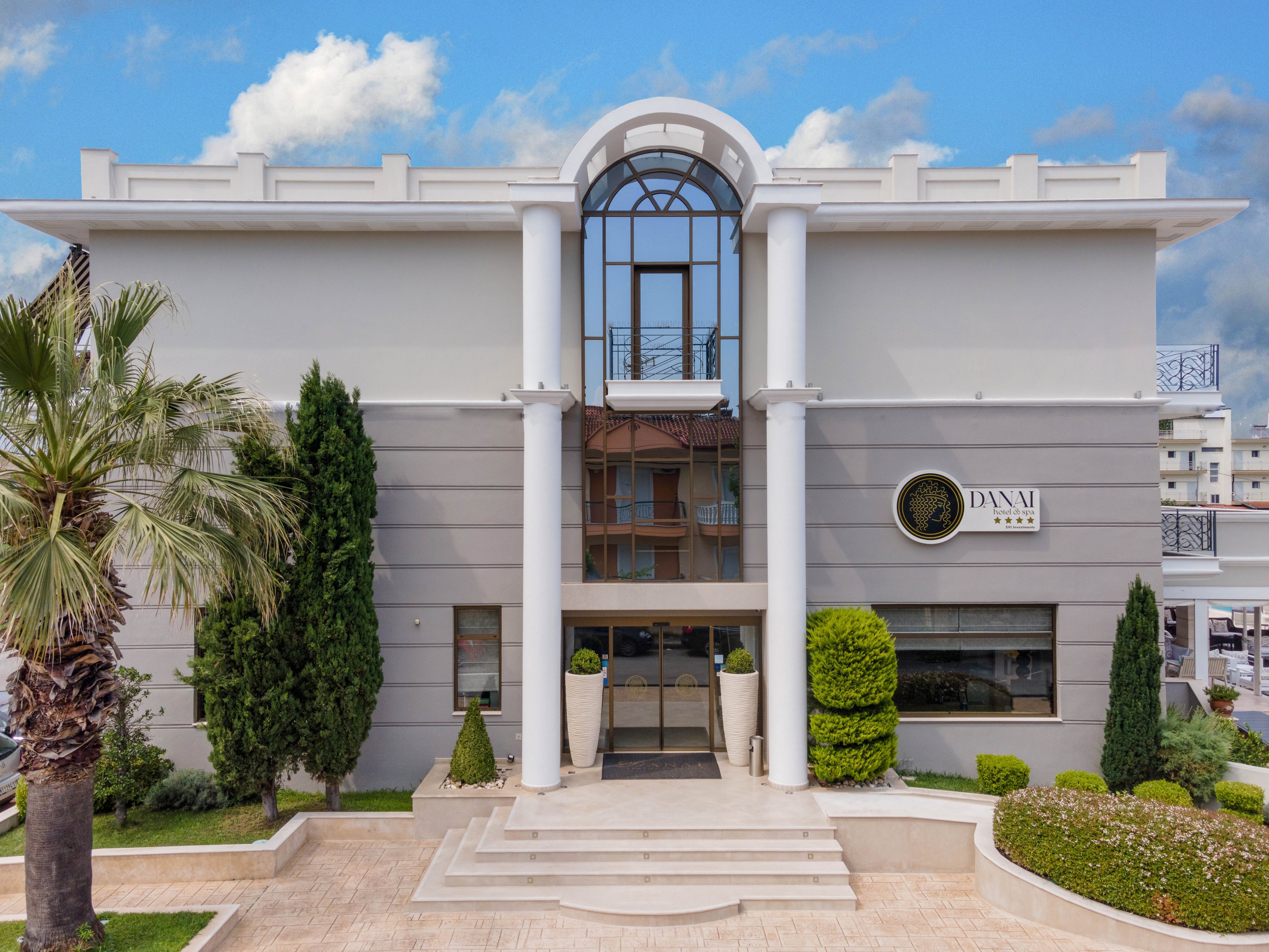 A contemporary two-story office building features prominent white columns and a glass archway above the entrance. The facade is accented by manicured greenery, including palm trees and shrubs, and a circular sign displaying the text 'DYNAMI'. The setting is urban, with blue skies and neighboring structures visible in the background. The overall mood is professional and inviting.
