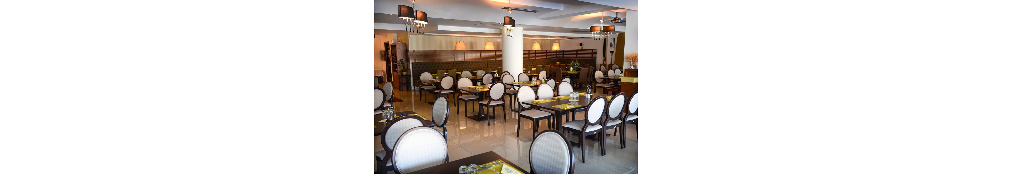 The image shows a spacious restaurant interior featuring neatly arranged tables and chairs with round backs. The setting is well-lit with warm overhead lamps and natural light, creating a welcoming atmosphere. The decor is contemporary, with neutral tones and polished floors, and the tables are set with placemats and glassware. No visible text or numeric values are present in the scene.
