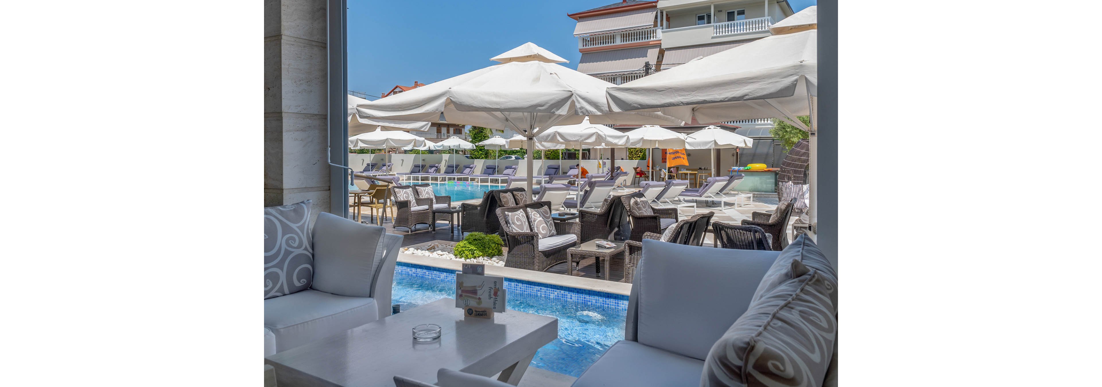 The image features a stylish outdoor lounge area with cushioned seating and a view of a swimming pool. Multiple wicker chairs and tables are arranged under large white sun umbrellas, creating a relaxing atmosphere. The setting is bright and sunny, with modern buildings in the background and a clear blue sky overhead. No visible text or numbers are present in the scene.