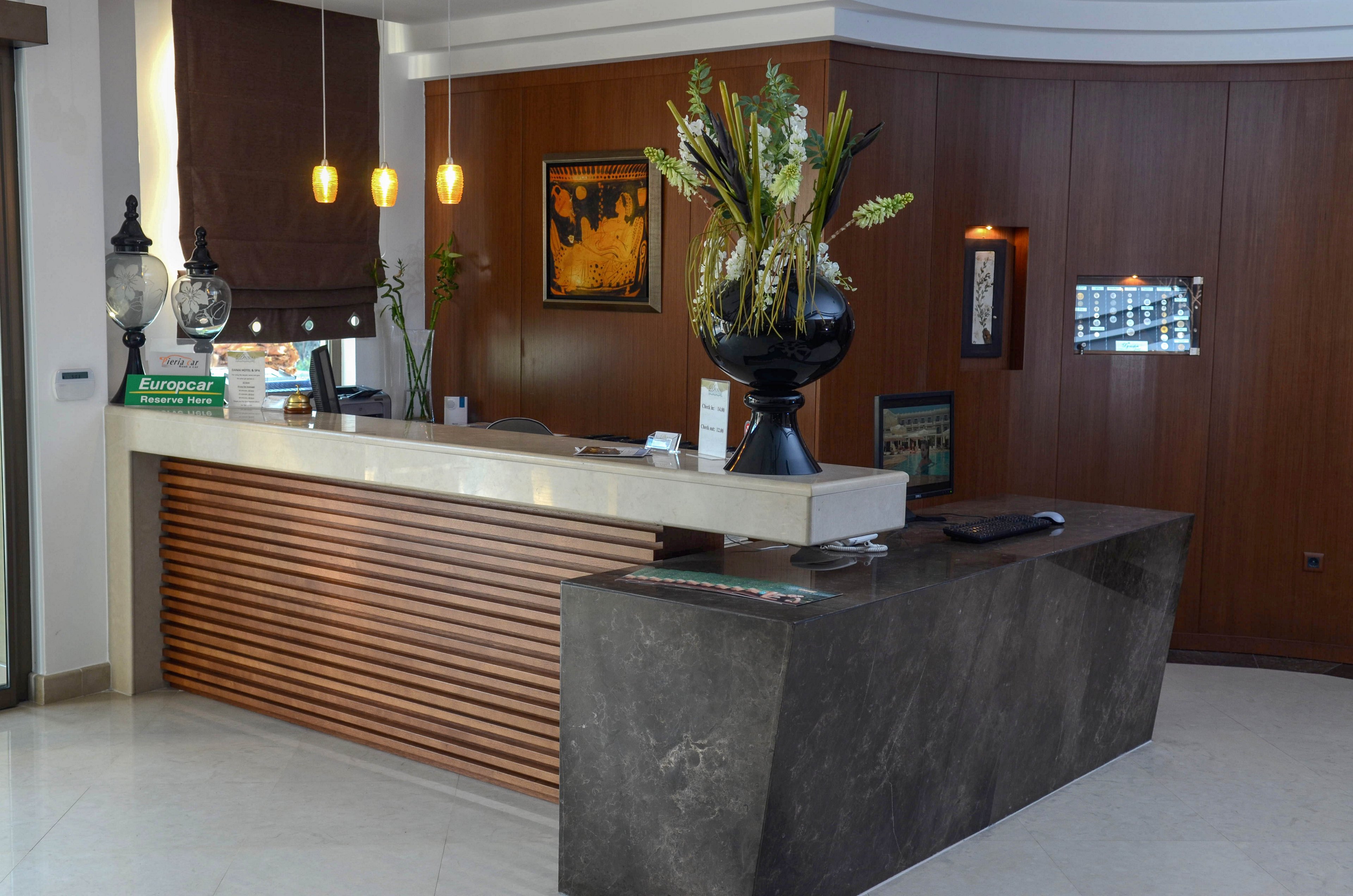 A contemporary hotel reception area features a sleek marble counter and a wooden accent wall. The desk is decorated with a large black vase filled with assorted flowers and greenery. Pendant lights hang above the counter, and a framed artwork is visible on the wall. The sign on the desk reads 'Europcar,' indicating a car rental service presence.