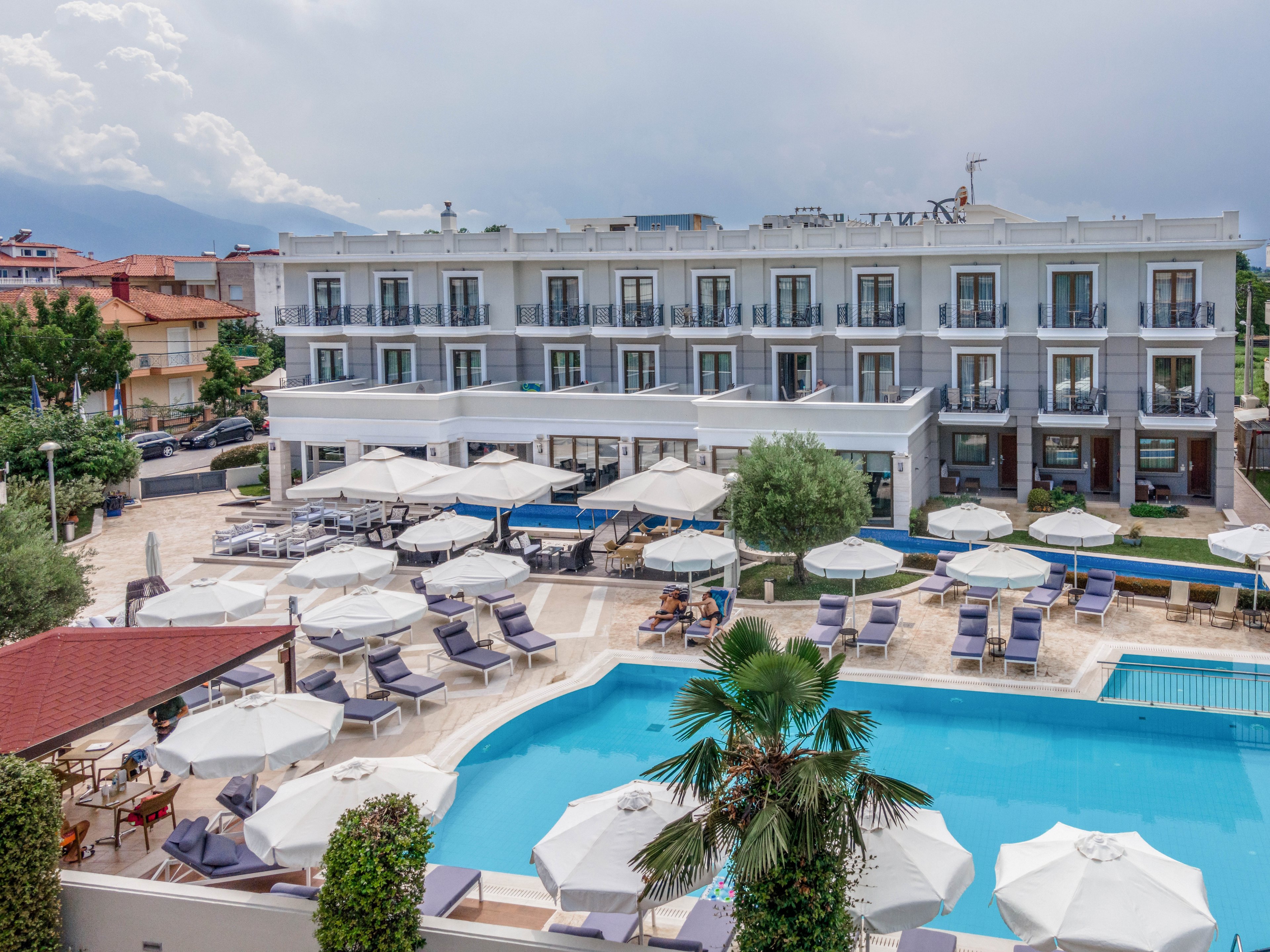 The image features a contemporary hotel building with multiple balconies overlooking a spacious outdoor pool area. Numerous lounge chairs and large white umbrellas surround the pool, creating a relaxing atmosphere. The setting appears to be a resort or vacation destination, with clear skies and landscaped greenery enhancing the scene. No visible text or numeric values are present in the image.