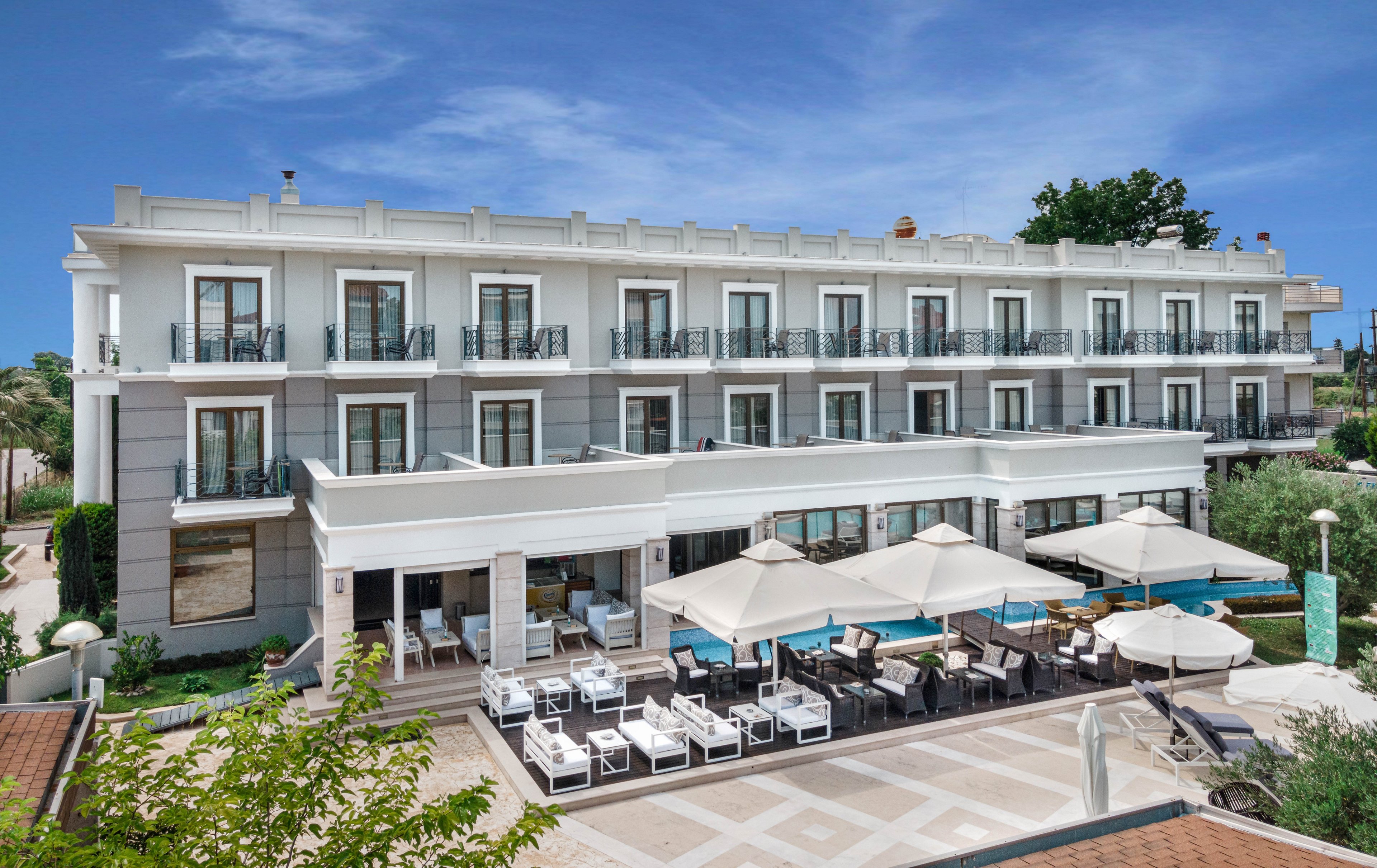 A contemporary multi-story hotel building is shown with balconies and large windows. The foreground features a poolside area with white lounge chairs and umbrellas, creating a relaxing outdoor setting. The scene is bright and sunny, with clear blue skies and lush greenery surrounding the property. The architectural style is clean and modern, emphasizing comfort and leisure.