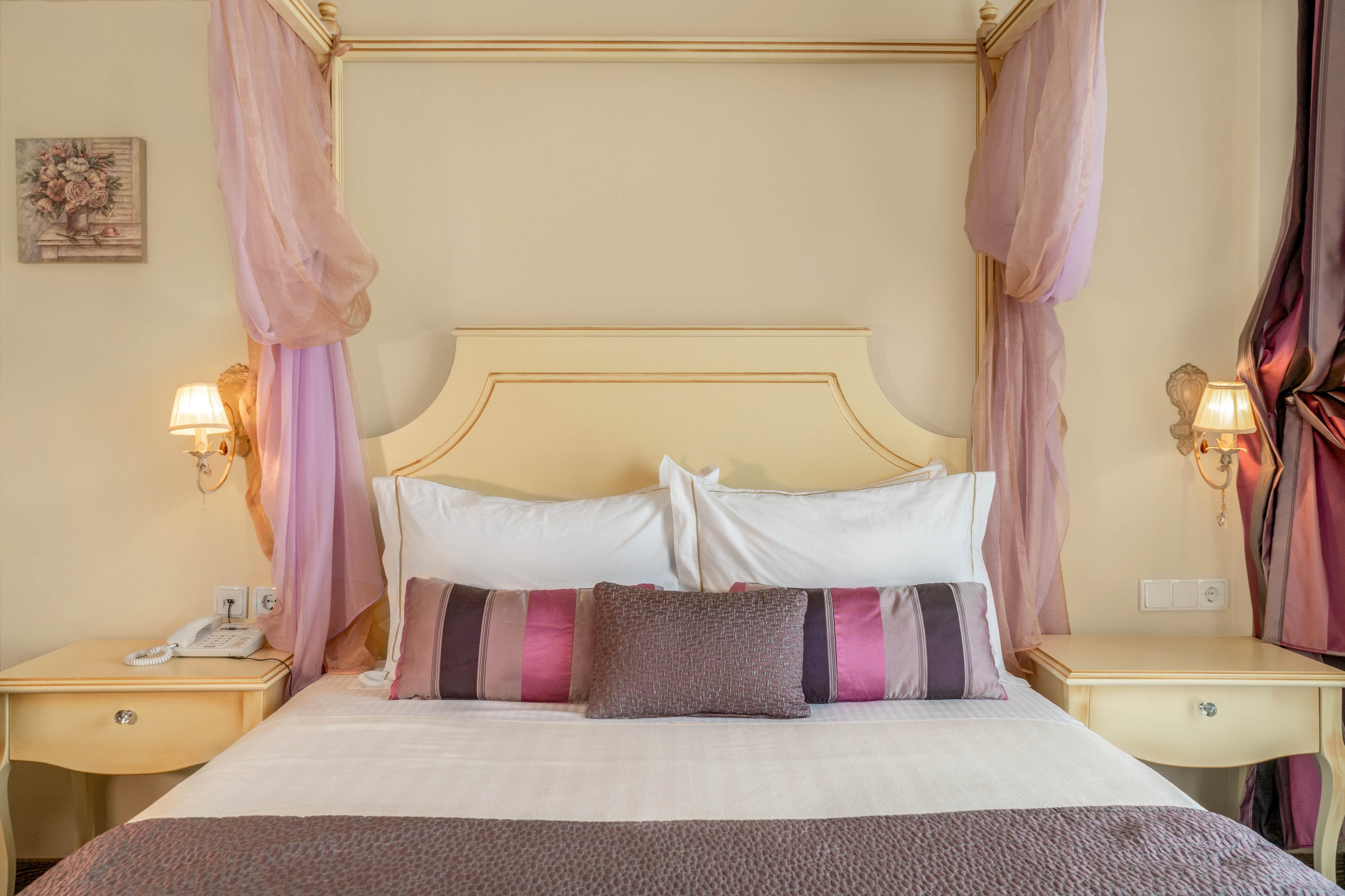 A neatly made bed with white linens and decorative pillows sits between two cream-colored nightstands in a softly lit hotel room. The headboard is framed by sheer pink drapes and wall-mounted lamps, creating a cozy and inviting atmosphere. The setting features pastel tones and subtle floral artwork, with a telephone and electrical outlets visible on the left nightstand.