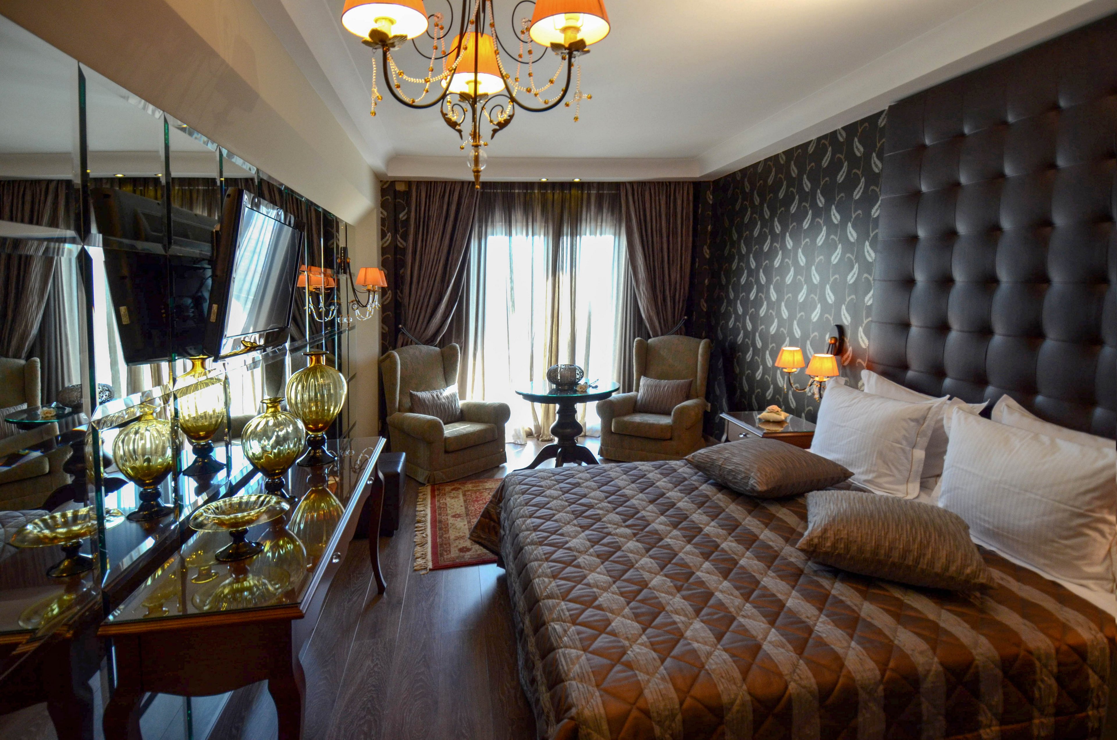 A stylish hotel room features a large bed with a quilted brown cover, two armchairs, and a small round table near the window. The space is decorated with dark tones, patterned wallpaper, and mirrored surfaces, creating a luxurious ambiance. Warm lighting from lamps and a chandelier enhances the cozy mood, while natural light filters through sheer curtains.