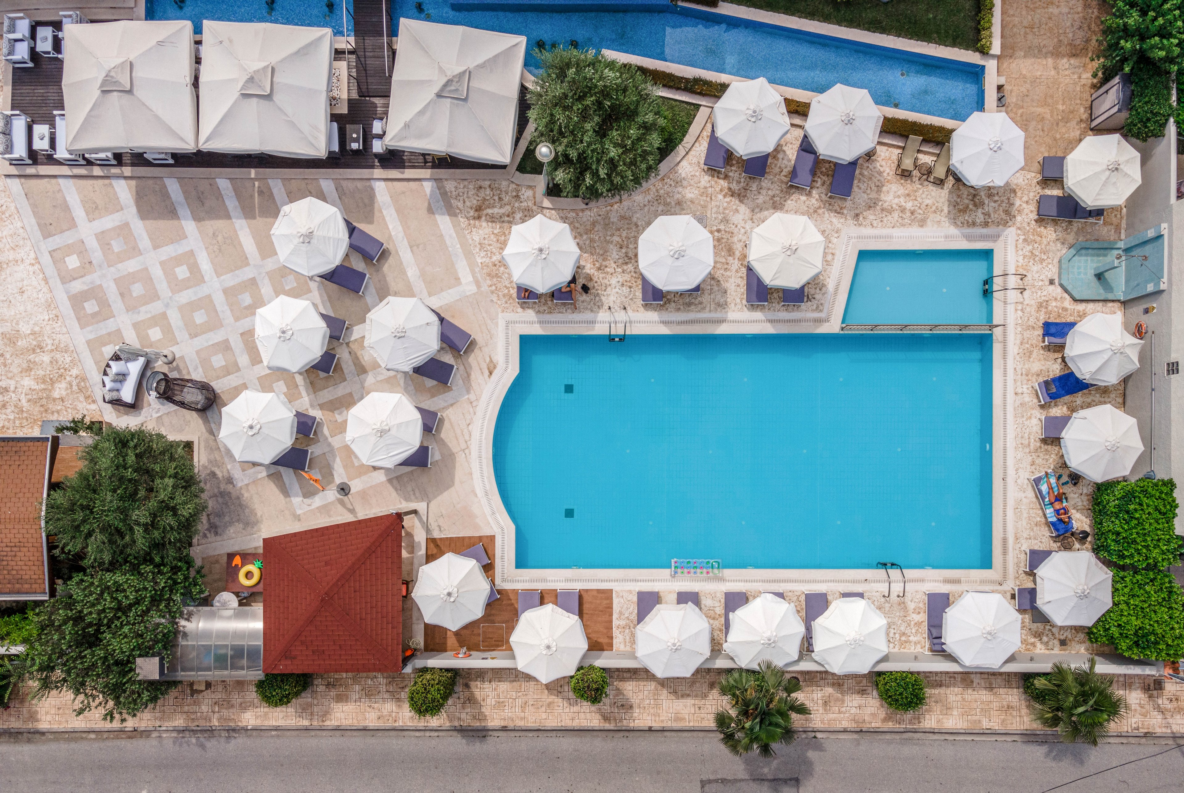 The main subject is a rectangular swimming pool surrounded by numerous white umbrellas, viewed from above. The setting appears to be a resort or hotel outdoor area, with poolside seating and cabanas visible. The visuals include clear blue water, geometric tile patterns, and lush greenery. No visible text or numbers are present in the image.