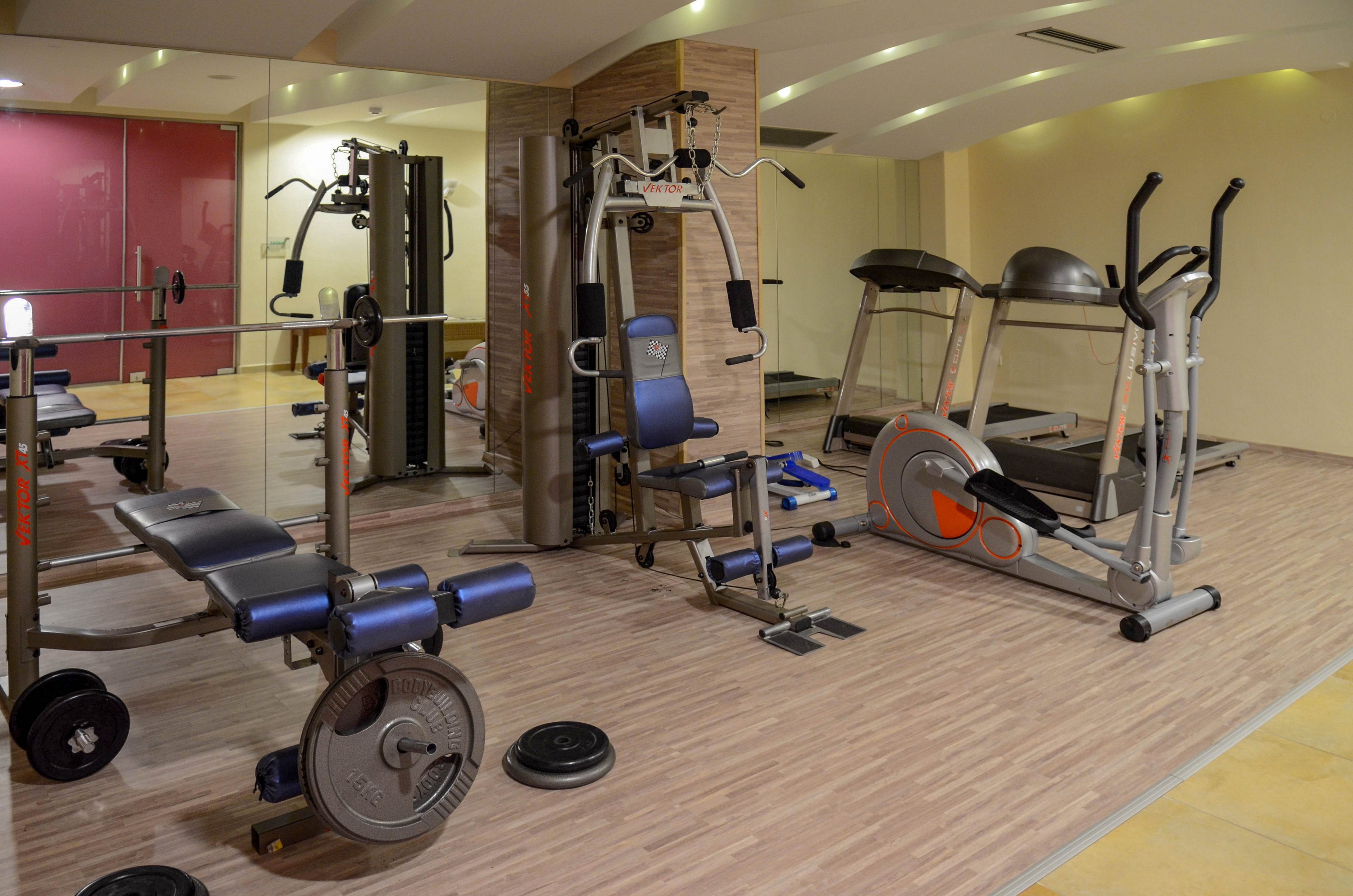 A contemporary gym space featuring various exercise machines, including a treadmill, stationary bike, and weight bench. The setting is indoors with wood-style flooring and mirrored walls, creating a clean and organized environment. The equipment is arranged for easy access, and the room is well-lit with neutral tones. No visible text or numeric values are present on the machines.