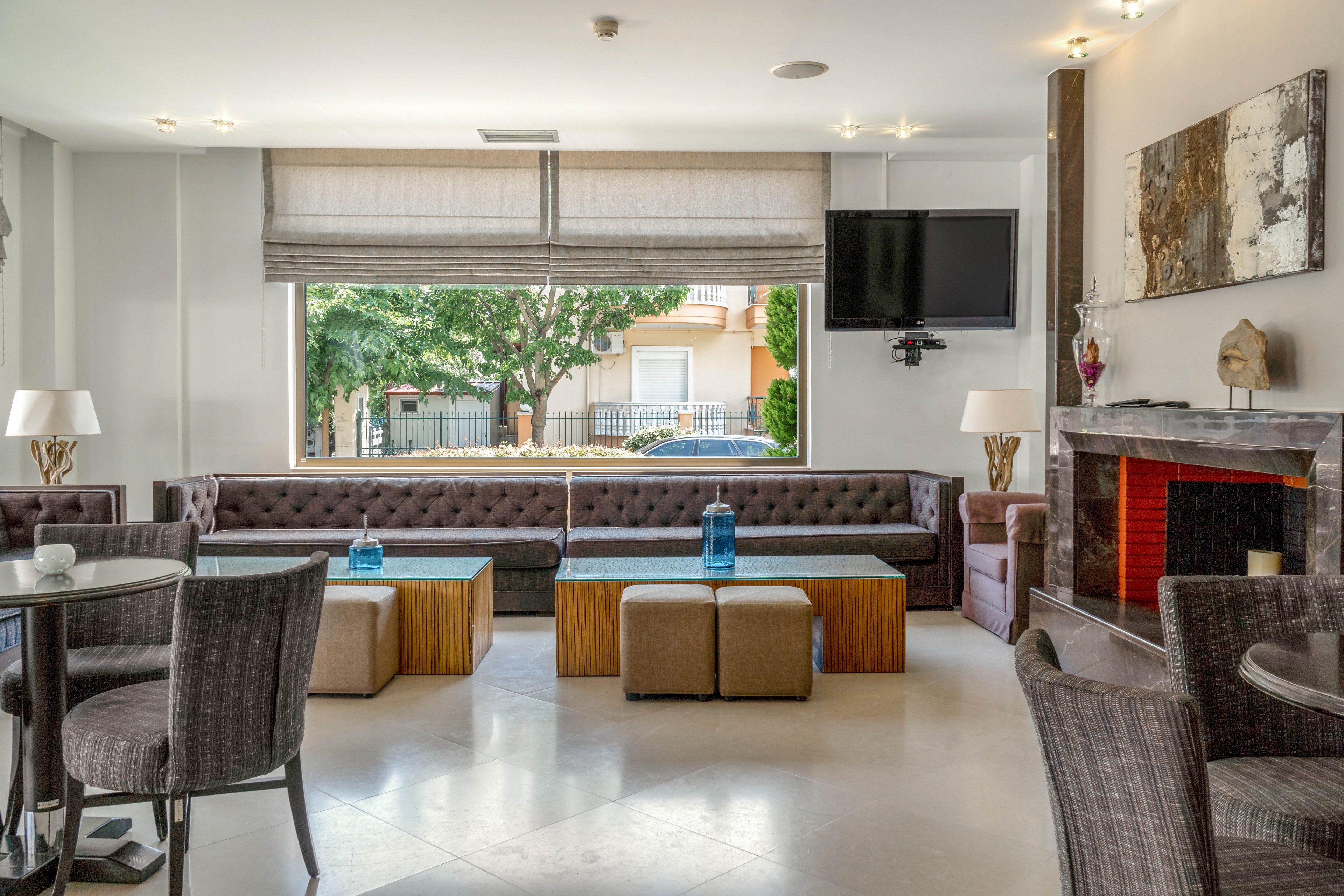 A contemporary lounge area features a long tufted sofa, two blue glass vases on wooden coffee tables, and several cushioned stools. The setting includes a fireplace, wall-mounted television, and large windows with a view of greenery and residential buildings outside. Neutral tones and soft lighting create a relaxed, inviting atmosphere, with abstract artwork and decorative lamps enhancing the stylish decor.
