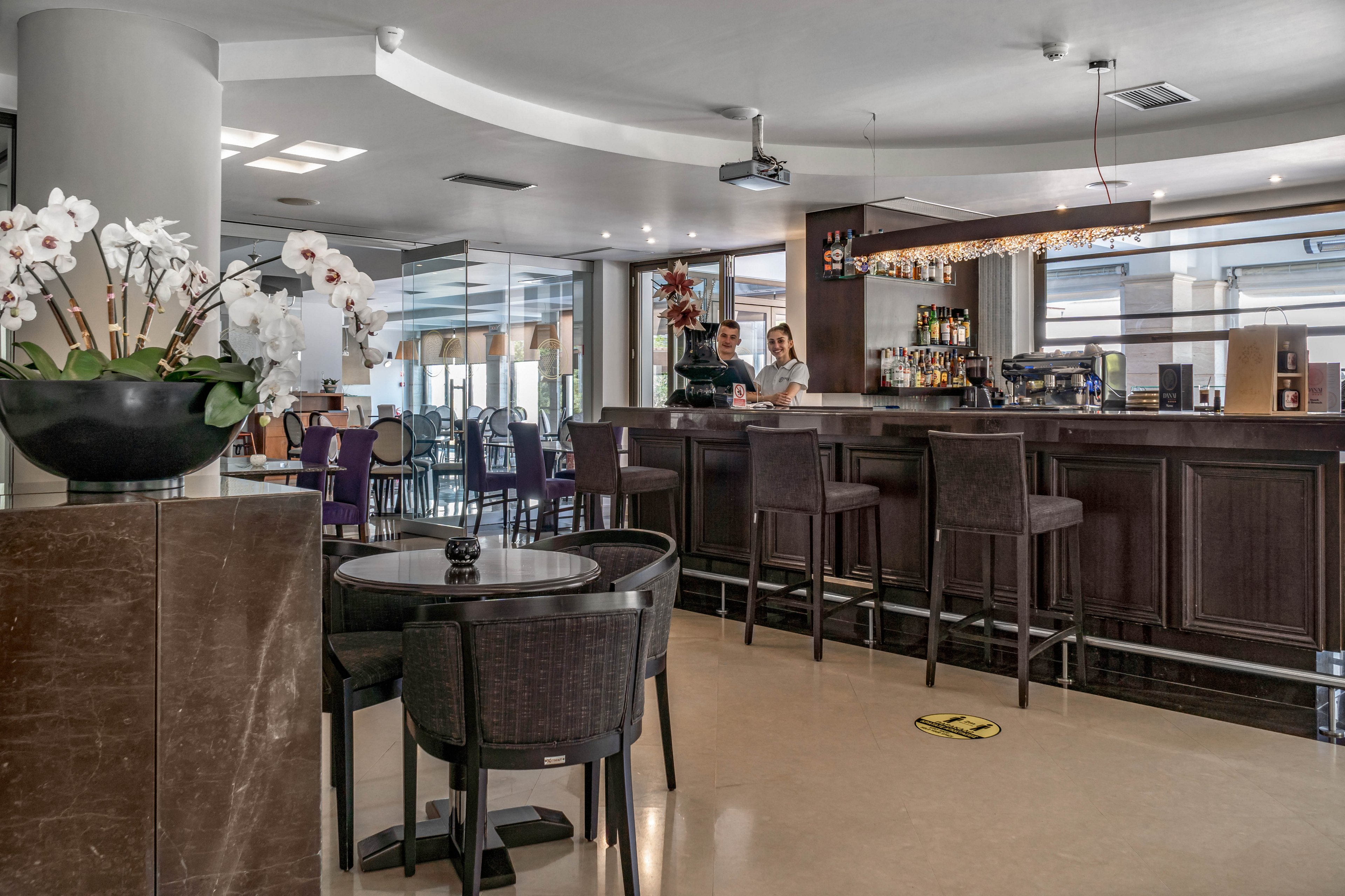A contemporary hotel bar features a dark wood counter with high chairs and a well-stocked back bar. Two staff members stand behind the counter, ready to serve guests. The setting is bright with natural light, and a large orchid arrangement decorates the foreground. A yellow floor marker with the number '1' is visible, indicating social distancing. The overall mood is welcoming and relaxed.