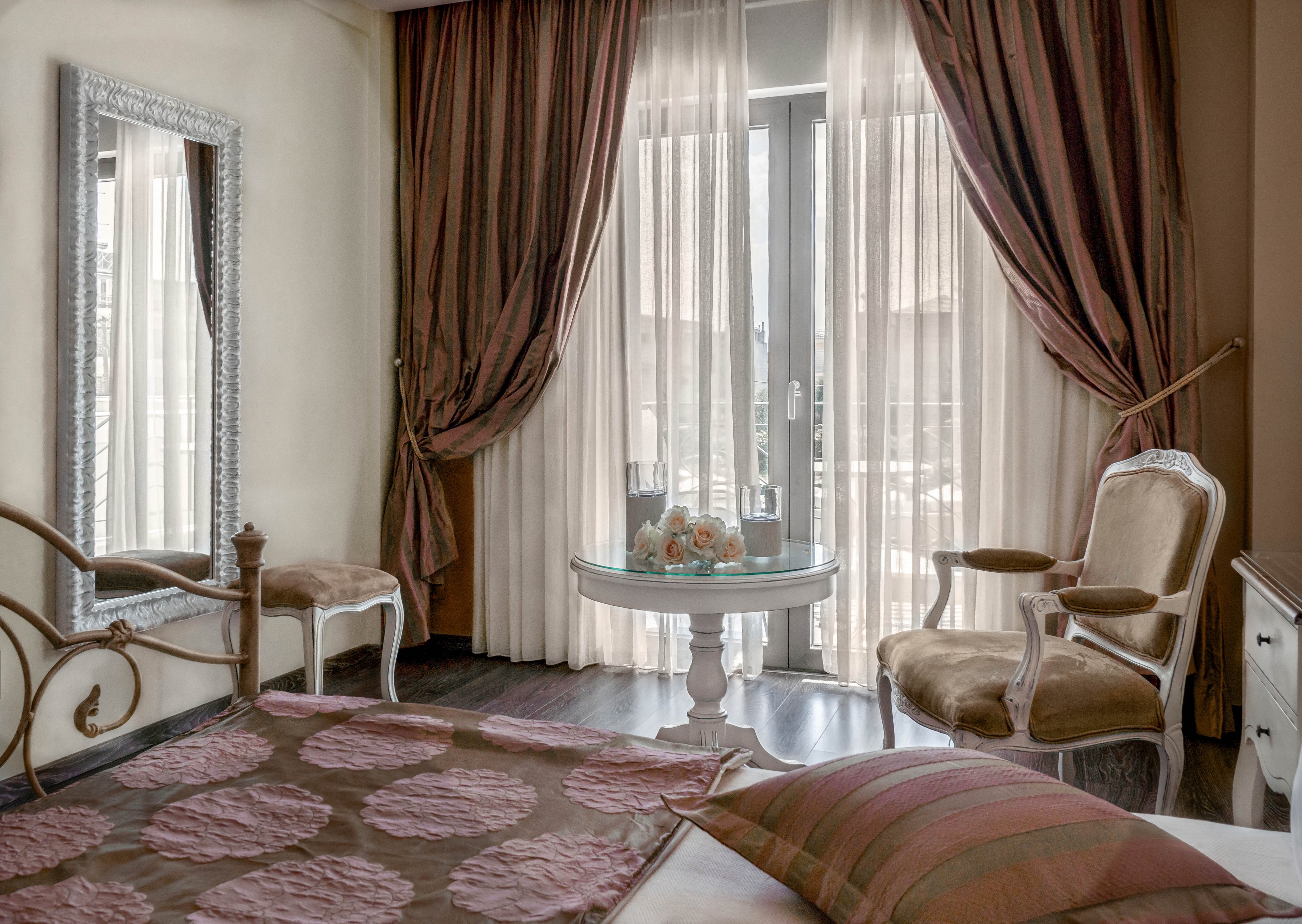 The image features a stylish bedroom with vintage-inspired furniture, including an ornate chair and a decorative mirror. Soft brown and beige tones dominate the palette, creating a warm and inviting mood. Sheer curtains allow natural light to filter in, highlighting a small round table with a floral arrangement. The bedspread and pillow display a subtle pattern, enhancing the classic ambiance.