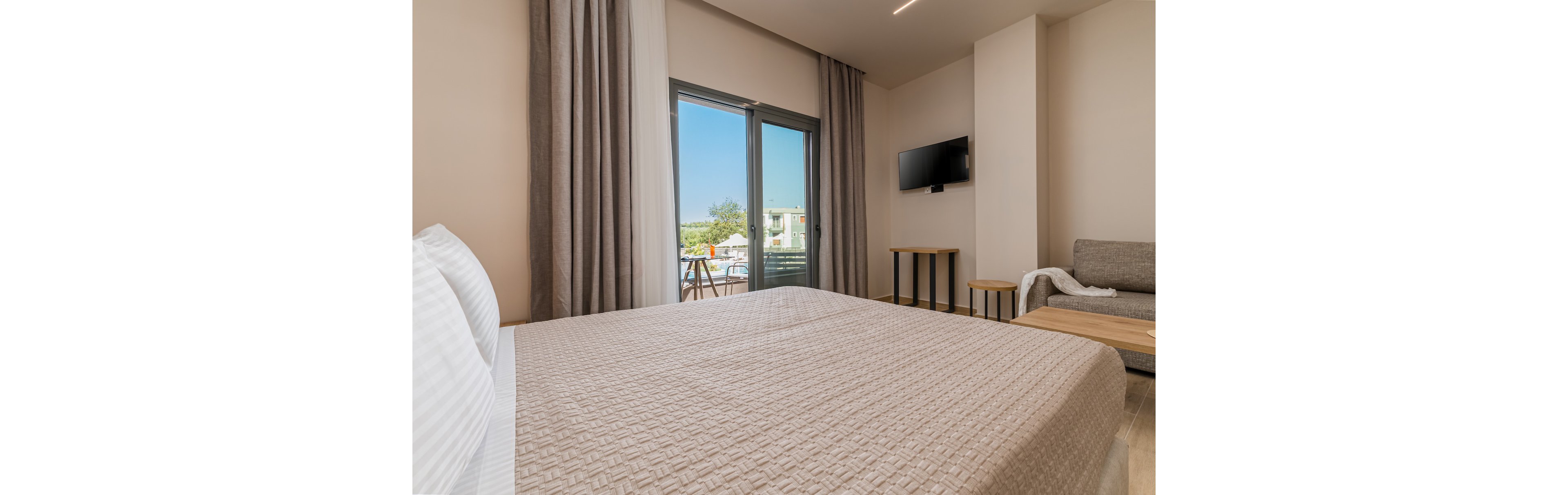 A neatly made double bed dominates a modern hotel room with neutral beige tones. Floor-to-ceiling curtains frame glass doors that open to a balcony overlooking a sunny outdoor scene. A wall-mounted flat-screen TV and small tables add functional comfort, while a light-colored sofa sits to the side. The overall atmosphere is bright, clean, and inviting, suitable for vacation or business travel.