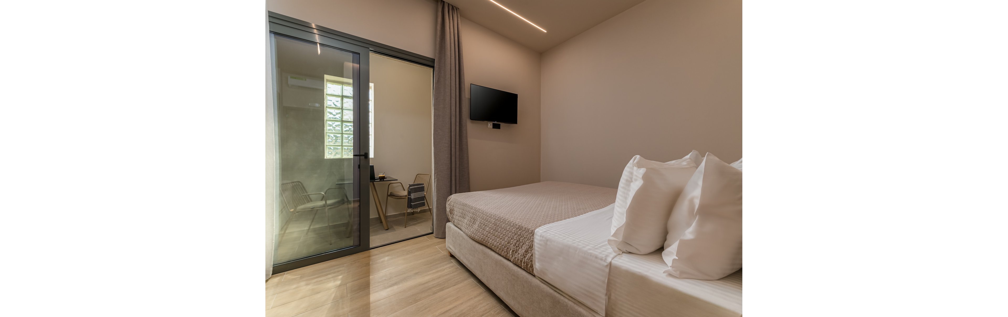 A contemporary bedroom features a neatly made double bed with layered pillows and a textured beige bedspread. A wall-mounted flat-screen TV faces the bed, and a floor-to-ceiling curtain frames a glass sliding door. Beyond the door, a small balcony or enclosed patio holds a table and chairs, with natural light streaming through a window. The space has a clean, minimalist design with neutral tones and smooth wooden flooring.