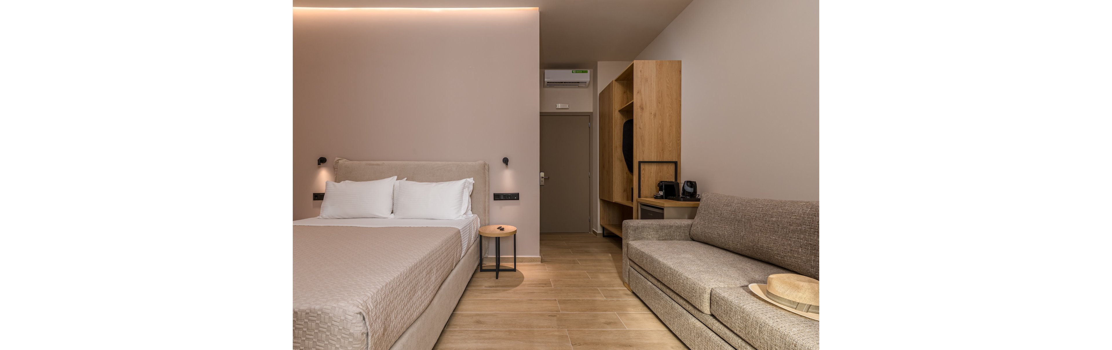A contemporary hotel room features a neatly made double bed with white pillows and beige bedding. Opposite the bed is a light-colored sofa with a straw hat resting on the seat. The space includes a wooden wardrobe unit with shelves, a round mirror, and a coffee machine, leading toward a closed door. Soft, warm lighting and neutral tones create a calm, minimalist atmosphere.