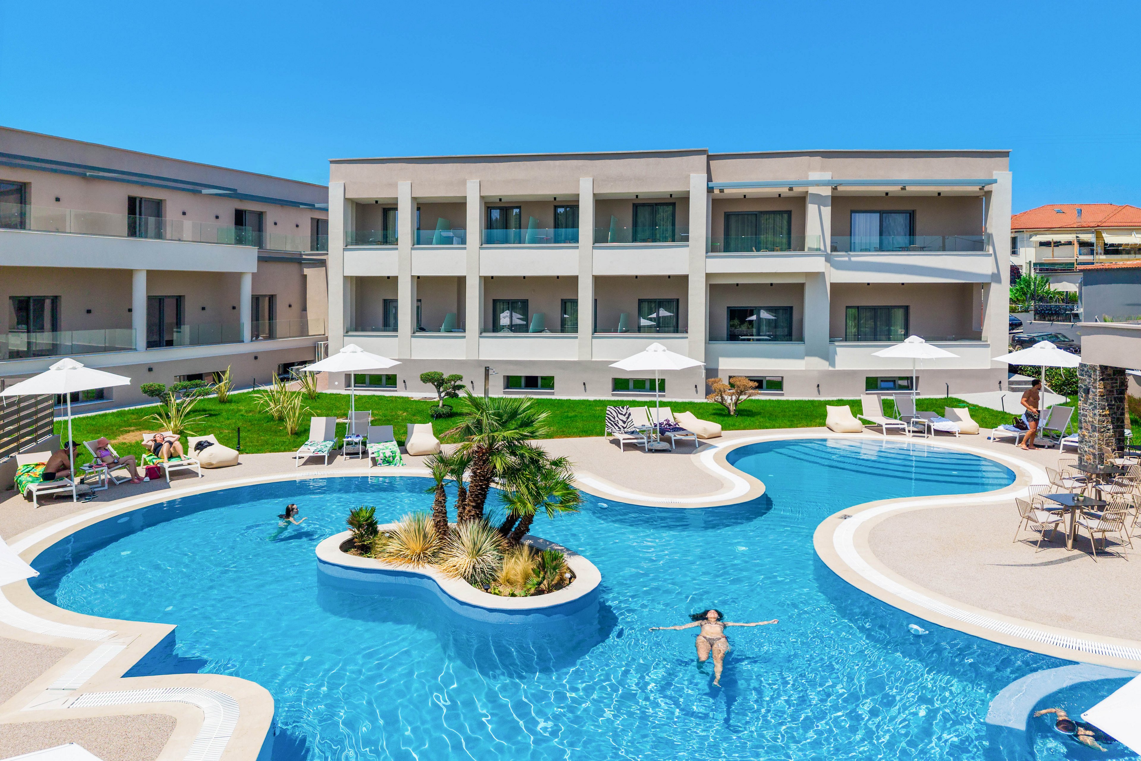 A contemporary three-story resort hotel overlooks a large curving outdoor swimming pool. Sun loungers and white umbrellas line the poolside terrace, with guests relaxing and swimming in the clear blue water. Palm trees and manicured lawns add a tropical touch to the courtyard setting. Bright sunlight and a clear blue sky create a vibrant, summery vacation atmosphere.