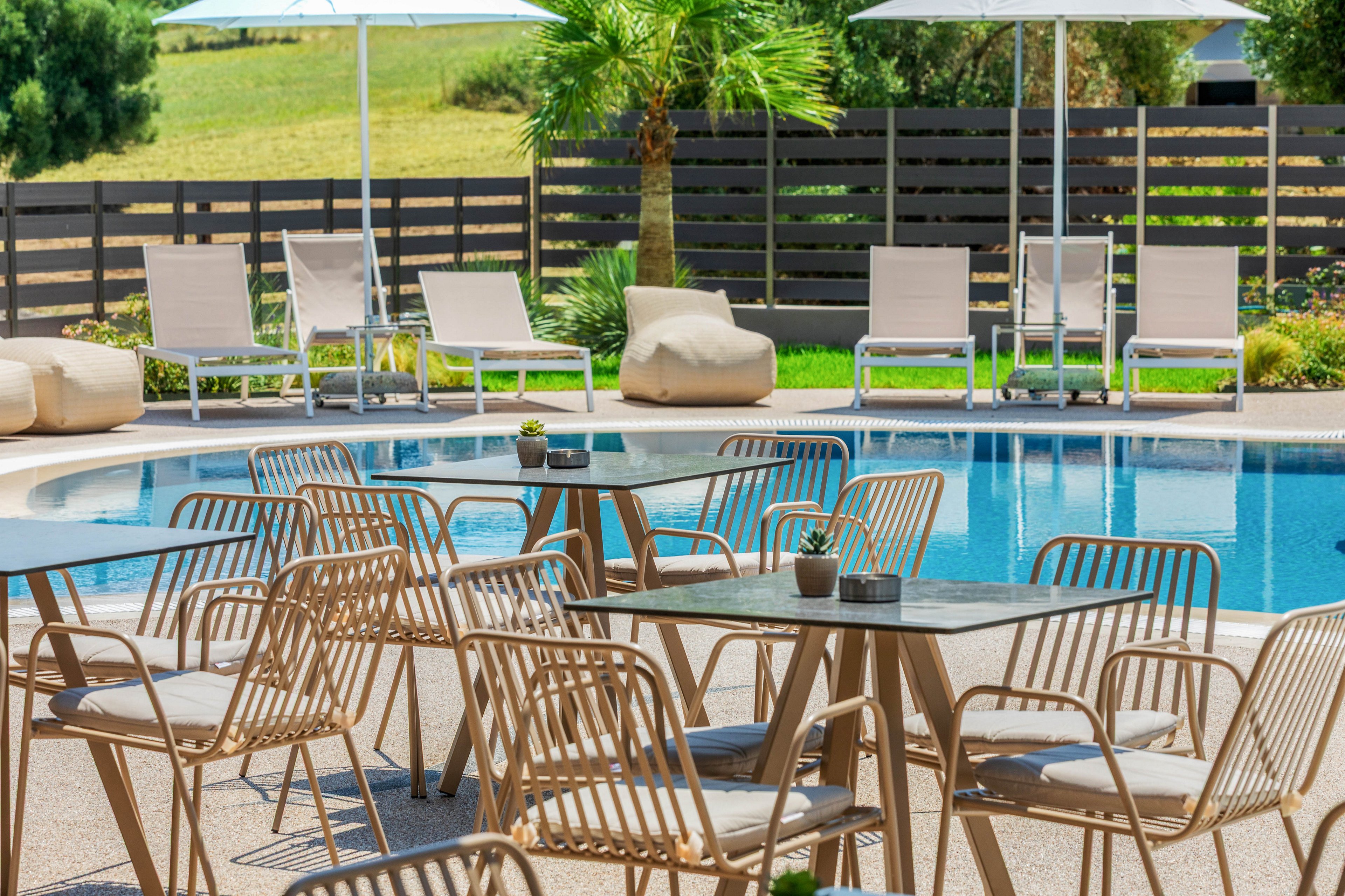 The image shows a contemporary outdoor pool area with several tables and chairs arranged on a sunlit terrace. In the background, cushioned loungers and umbrellas line the edge of the swimming pool, creating a relaxed resort atmosphere. A palm tree and manicured lawn add a touch of greenery, while a horizontal fence provides privacy. The overall scene suggests a comfortable, stylish space for leisure, dining, or social gatherings.