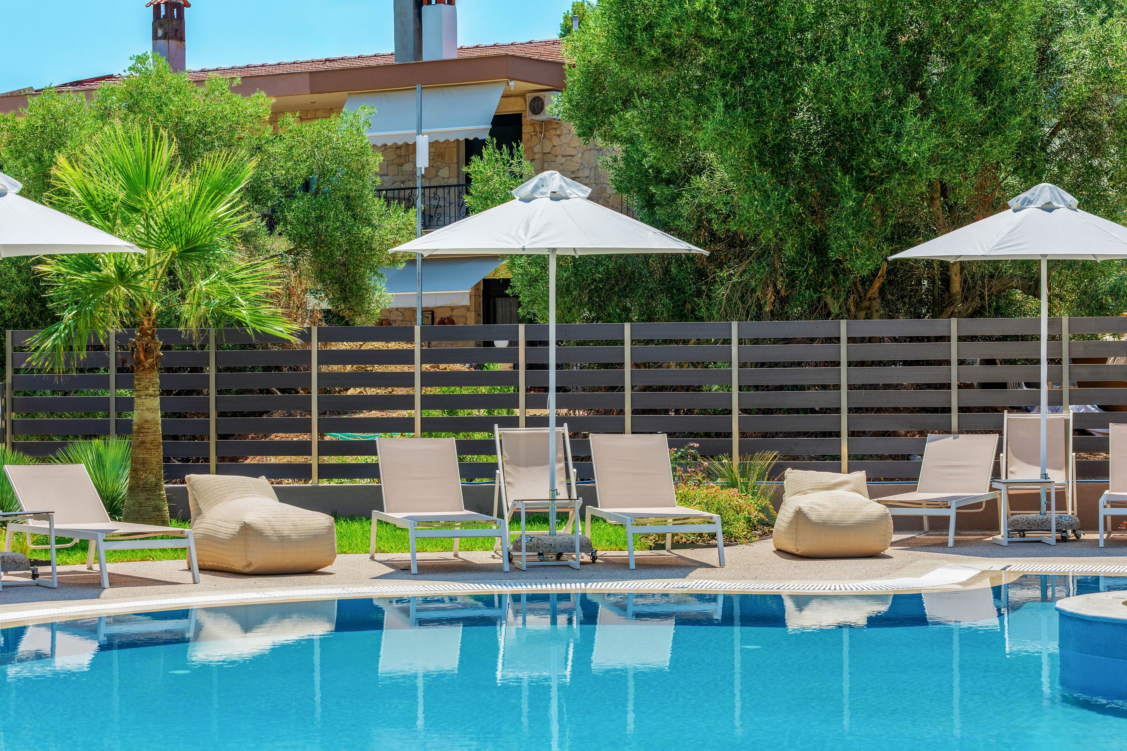 An inviting outdoor swimming pool is surrounded by beige sun loungers and cushioned seats under white umbrellas. A wooden fence and lush green trees create a private, tranquil backdrop. A palm tree adds a tropical touch to the bright, sunny setting. The scene suggests a relaxing vacation atmosphere at a modern resort.