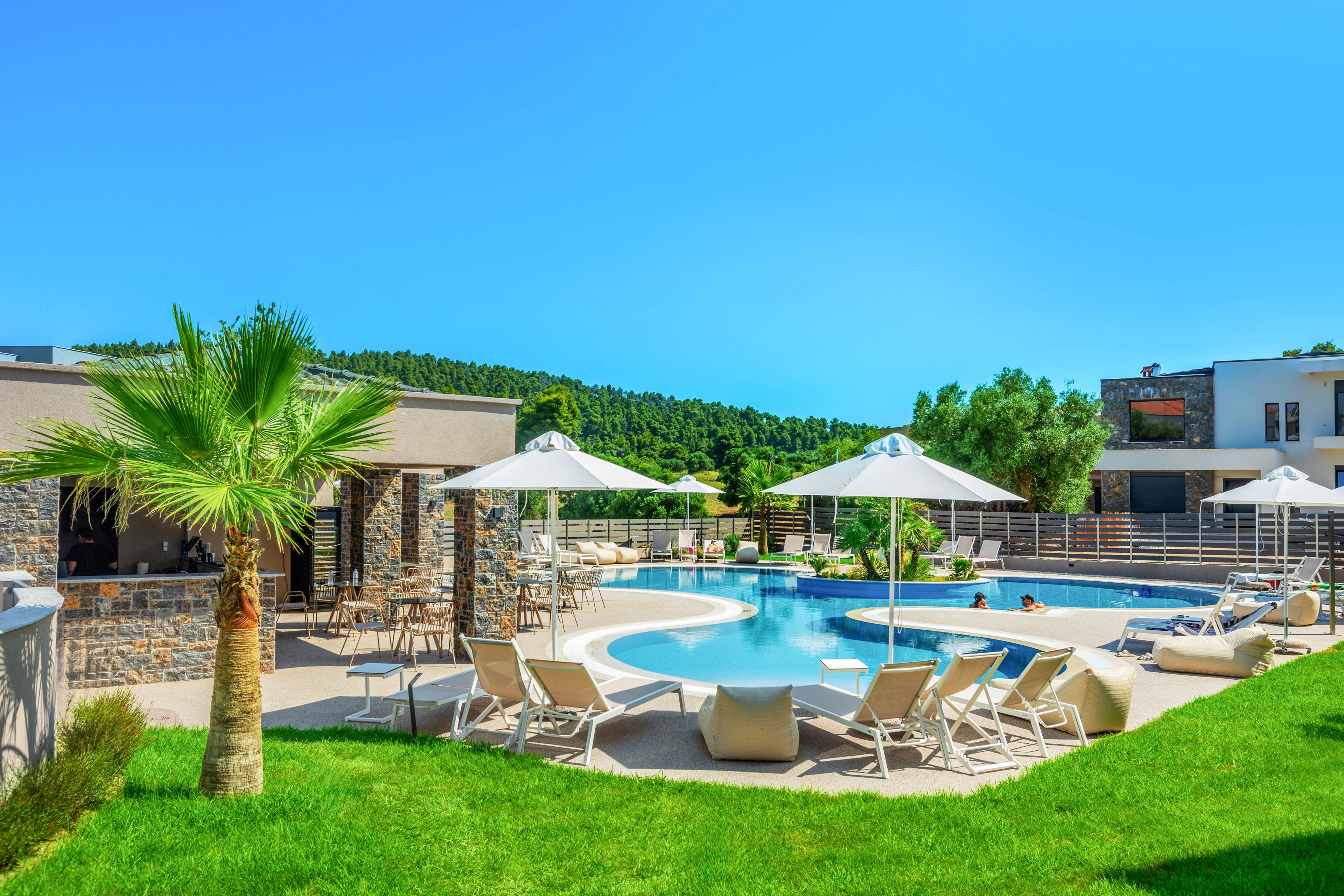 A modern resort-style swimming pool is surrounded by white sun loungers and umbrellas on a bright sunny day. Contemporary low-rise buildings with stone and concrete facades frame the pool area. A neatly manicured lawn with a small palm tree adds a tropical touch in the foreground. In the background, lush green hills and clear blue sky create a relaxing vacation atmosphere.