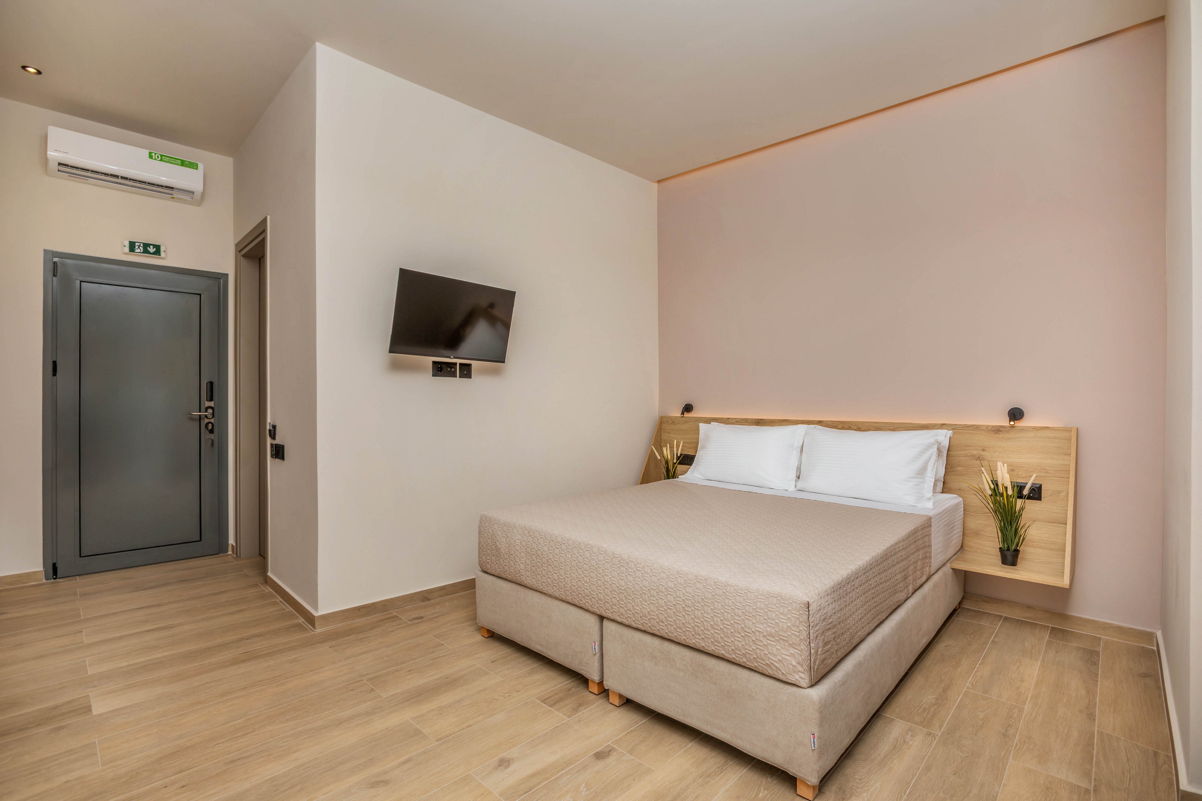 A contemporary hotel bedroom features a double bed with neutral bedding and two white pillows against a light wooden headboard. The room has light-colored walls and wood-look flooring, creating a clean and minimalist atmosphere. A flat-screen TV is mounted on the wall opposite the bed, and a gray door and air conditioning unit are visible. Small decorative plants add a touch of greenery to the simple interior design.