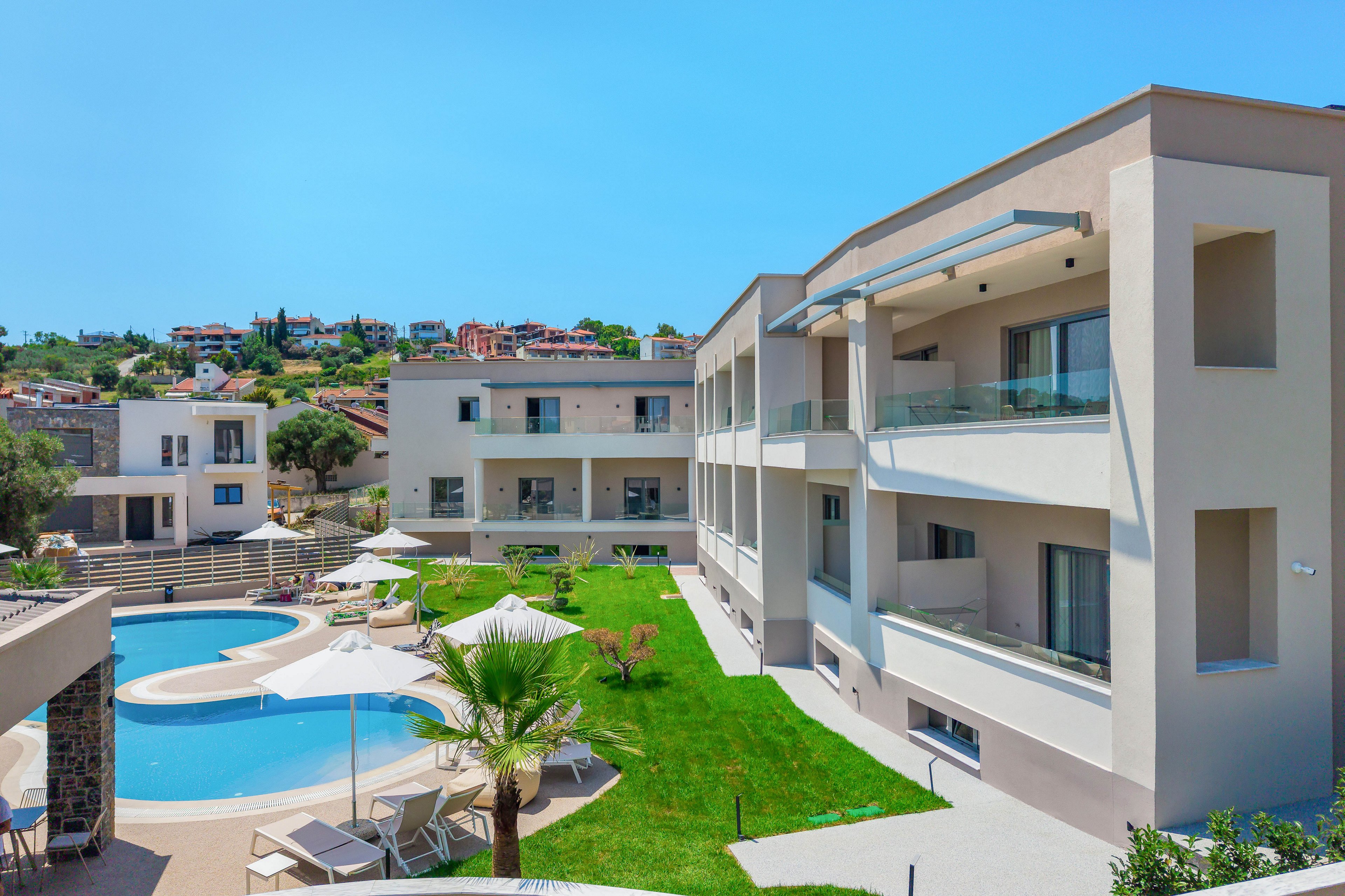 Contemporary low-rise apartment buildings surround a curved outdoor swimming pool in a sunny resort setting. The scene features manicured green lawns, palm trees, and poolside lounge chairs with umbrellas. Glass balconies and clean architectural lines emphasize a modern, minimalist style. In the background, additional houses and buildings rise on a gentle hillside under a clear blue sky.