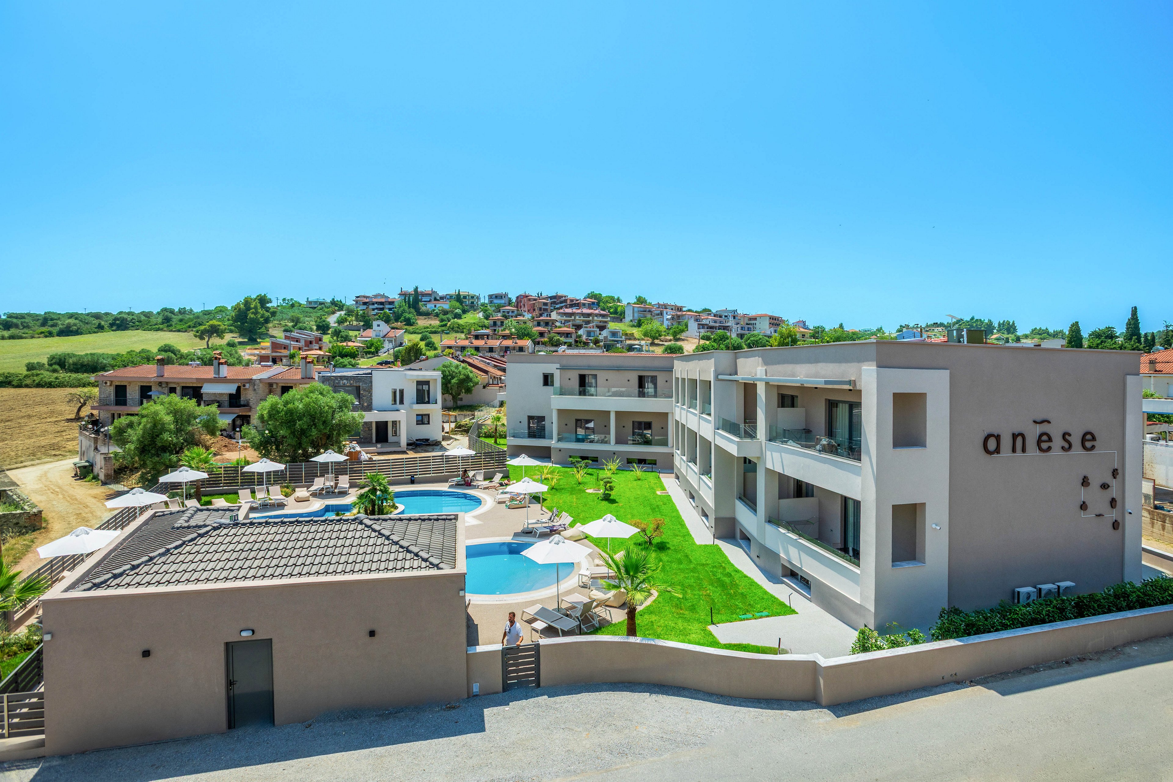 A contemporary resort complex named anēse is shown under a clear blue sky, featuring minimalist light-colored buildings and balconies. At the center of the property is a circular swimming pool surrounded by sun loungers and umbrellas on neatly maintained green lawns. The resort is set on the edge of a small town with houses and rolling fields visible in the background, suggesting a tranquil vacation destination. The overall scene conveys a bright, sunny atmosphere ideal for relaxation and leisure.