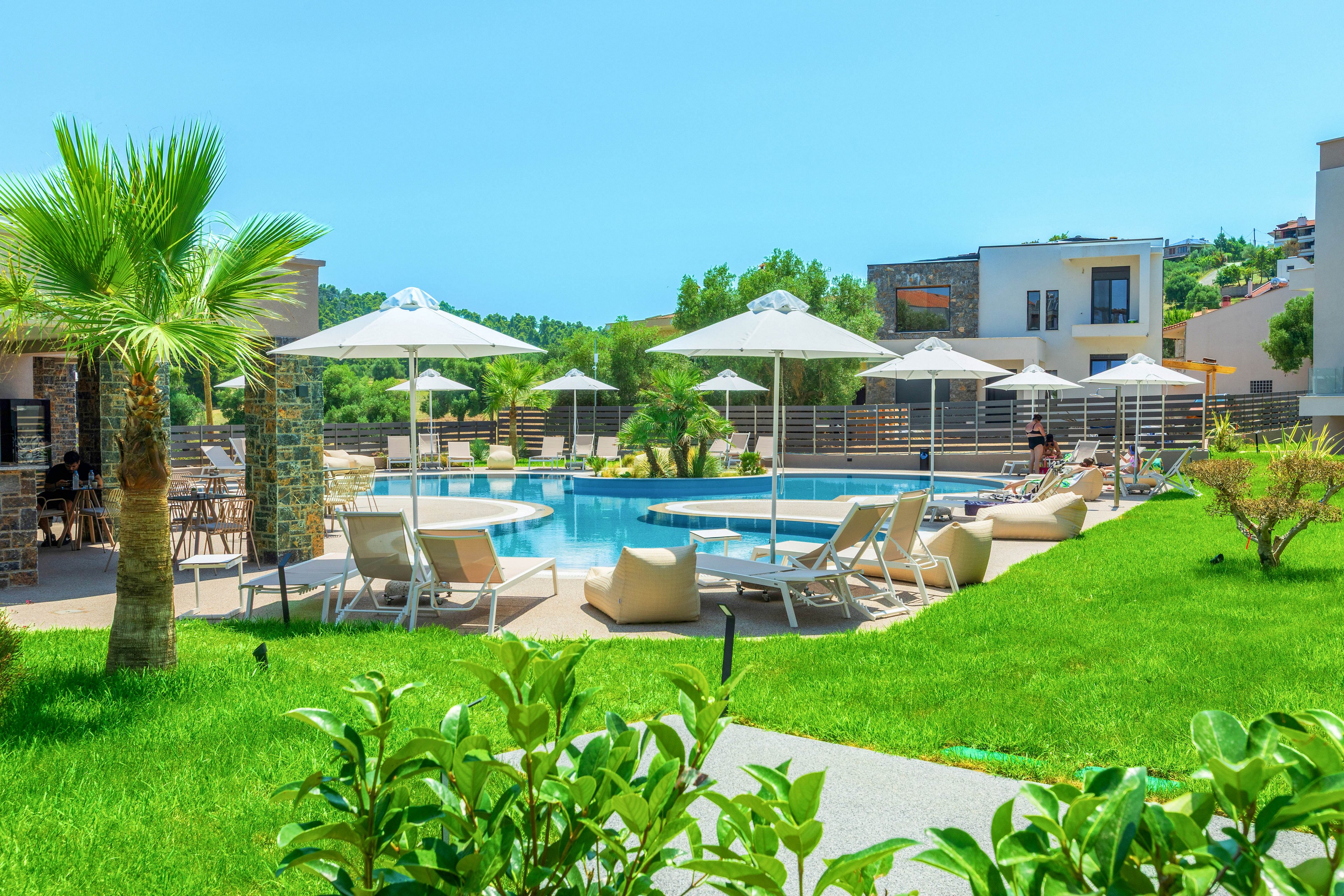 A contemporary resort pool area is surrounded by lush green lawns and tropical plants. White sun loungers and large umbrellas line the poolside, creating a relaxing vacation atmosphere. Modern low-rise buildings frame the scene under a clear blue sky. The overall setting suggests a sunny, tranquil holiday destination.
