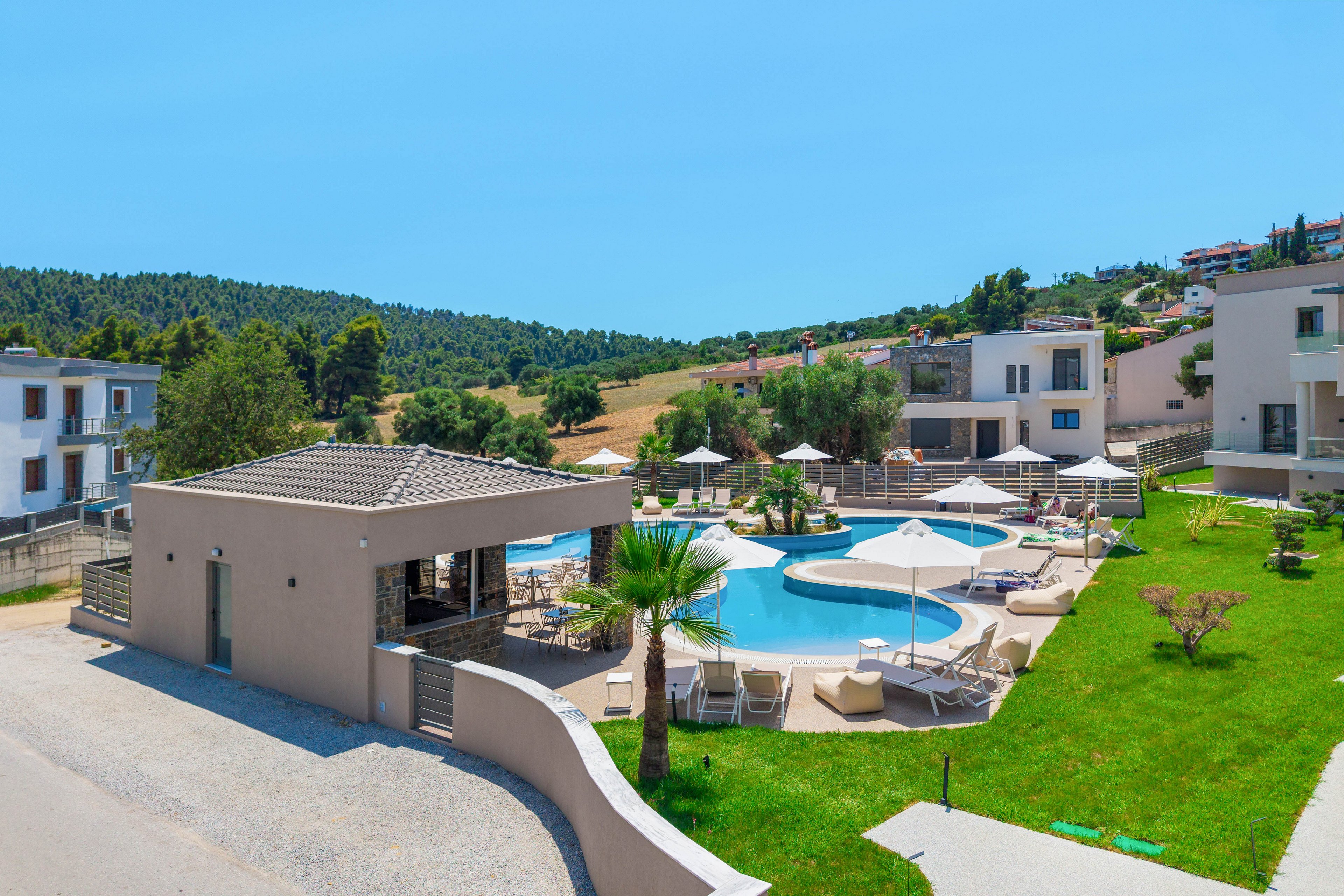A contemporary holiday resort features a curved outdoor swimming pool surrounded by white sun loungers and umbrellas. Low-rise modern buildings with balconies frame the pool area, set within neatly maintained green lawns. A small poolside bar or pavilion sits near the entrance, adjacent to a paved road. In the background, rolling hills and scattered houses create a peaceful countryside setting under a clear blue sky.