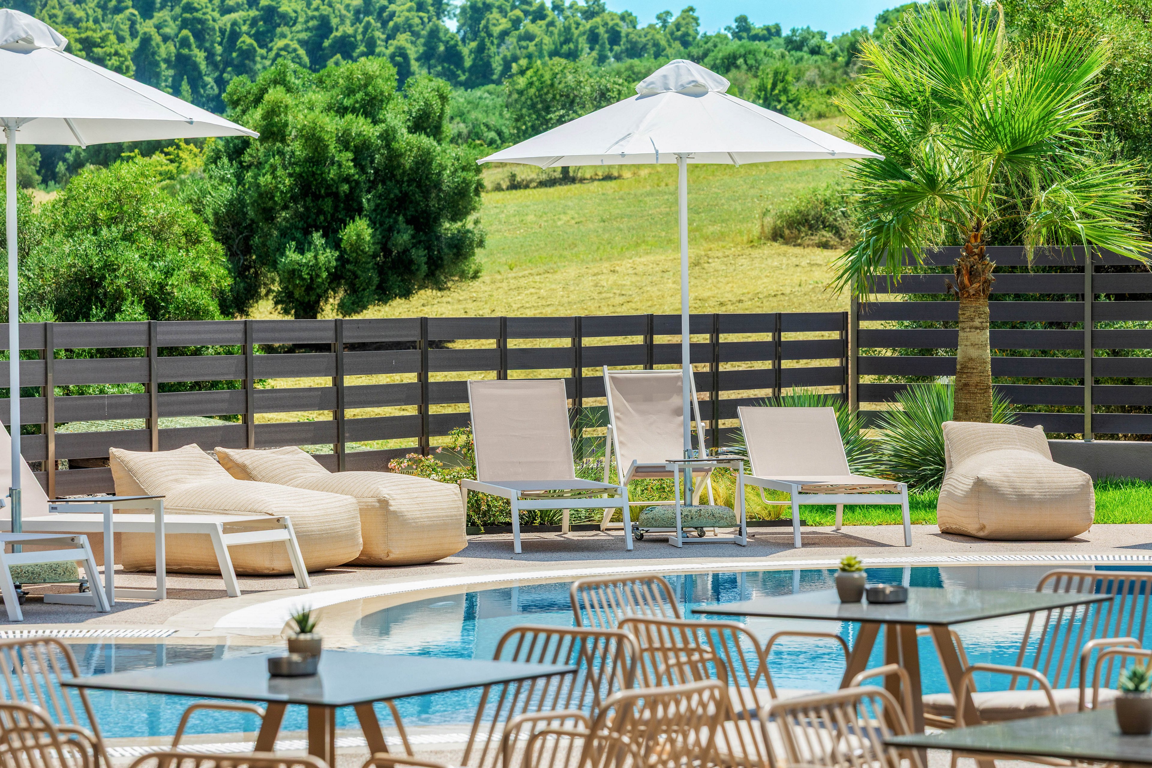 Outdoor resort pool area features modern lounge chairs and cushioned beanbag seats arranged around a curved swimming pool. White sun umbrellas provide shade, while metal chairs and small tables with potted plants sit in the foreground. A wooden fence, palm tree, and lush green landscape create a relaxed vacation atmosphere. Bright daylight and clean contemporary furniture emphasize a fresh, inviting summer setting.