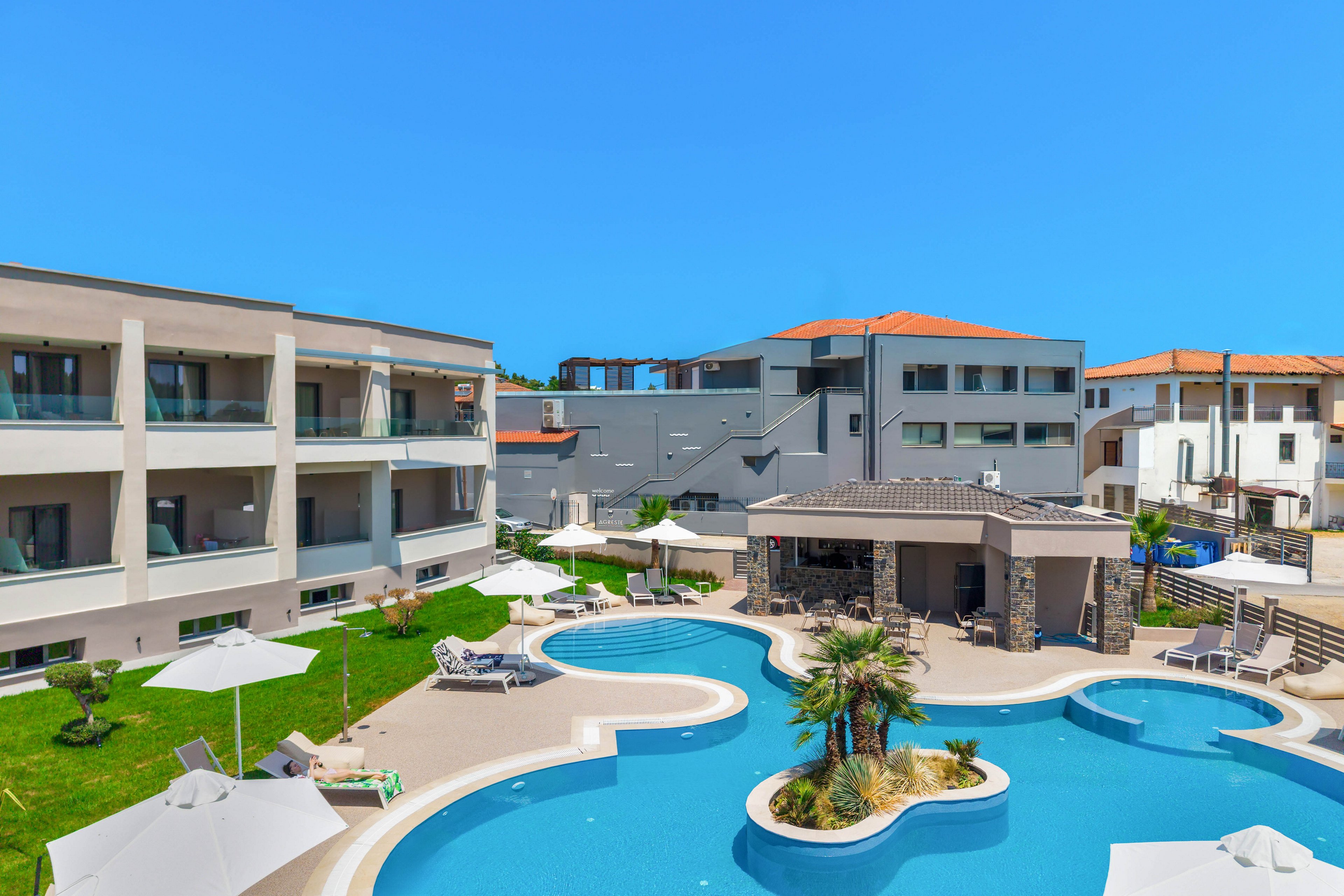 A contemporary resort hotel features a large, curving outdoor swimming pool at its center. The pool includes rounded sections, a small island with palm trees, and a swim-up bar or shaded pavilion. Sun loungers and white umbrellas are arranged neatly around the pool on green lawns and tiled decks. The surrounding low-rise buildings have balconies and flat roofs, with a bright blue sky overhead suggesting a sunny, warm climate.