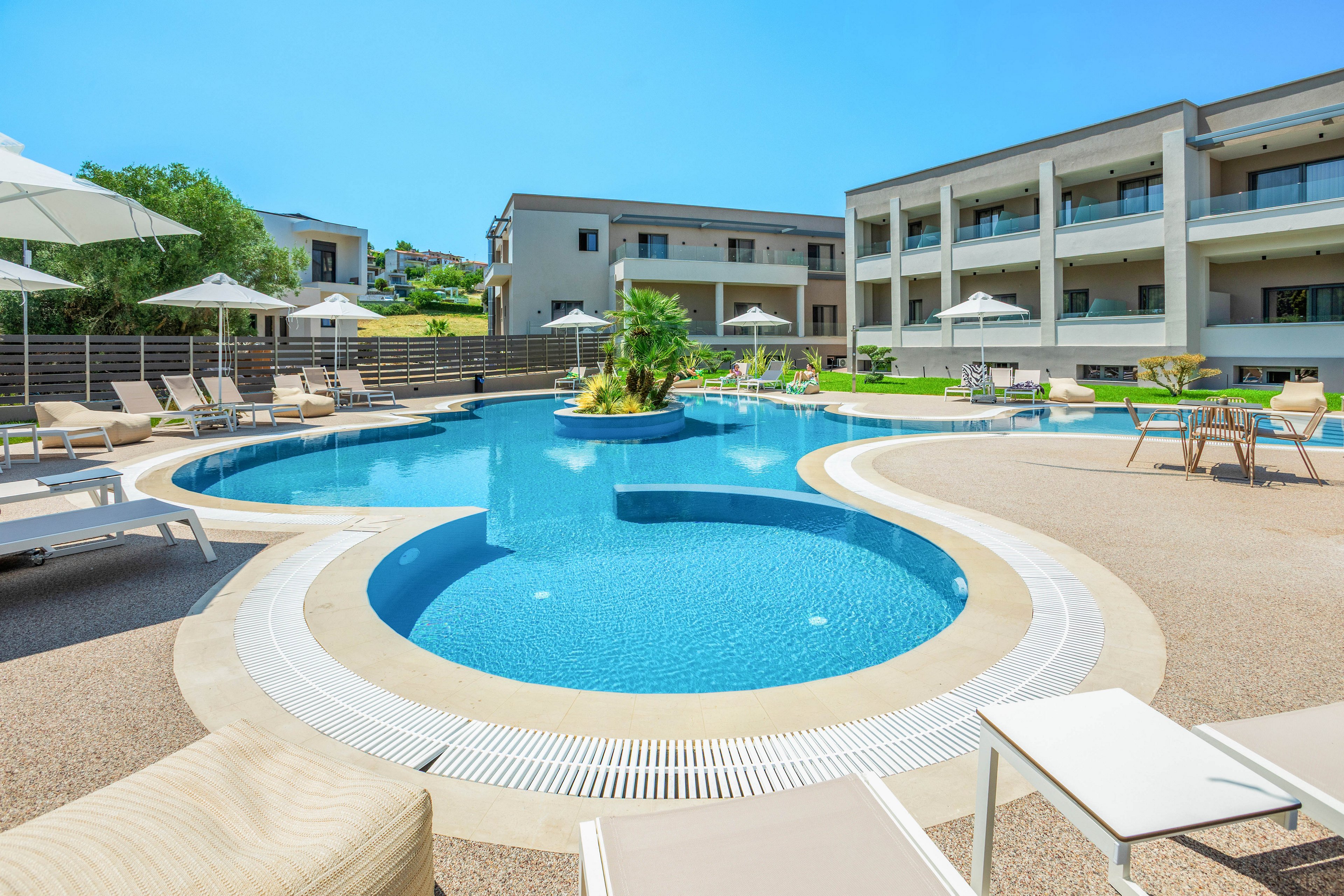 A contemporary resort features a curved outdoor swimming pool surrounded by sun loungers and umbrellas. The pool area is set within a clean, minimalist courtyard between low-rise hotel buildings. Bright blue water contrasts with the beige pool deck and neutral architecture under a clear sunny sky. The scene suggests a relaxing vacation atmosphere with ample seating and open space.