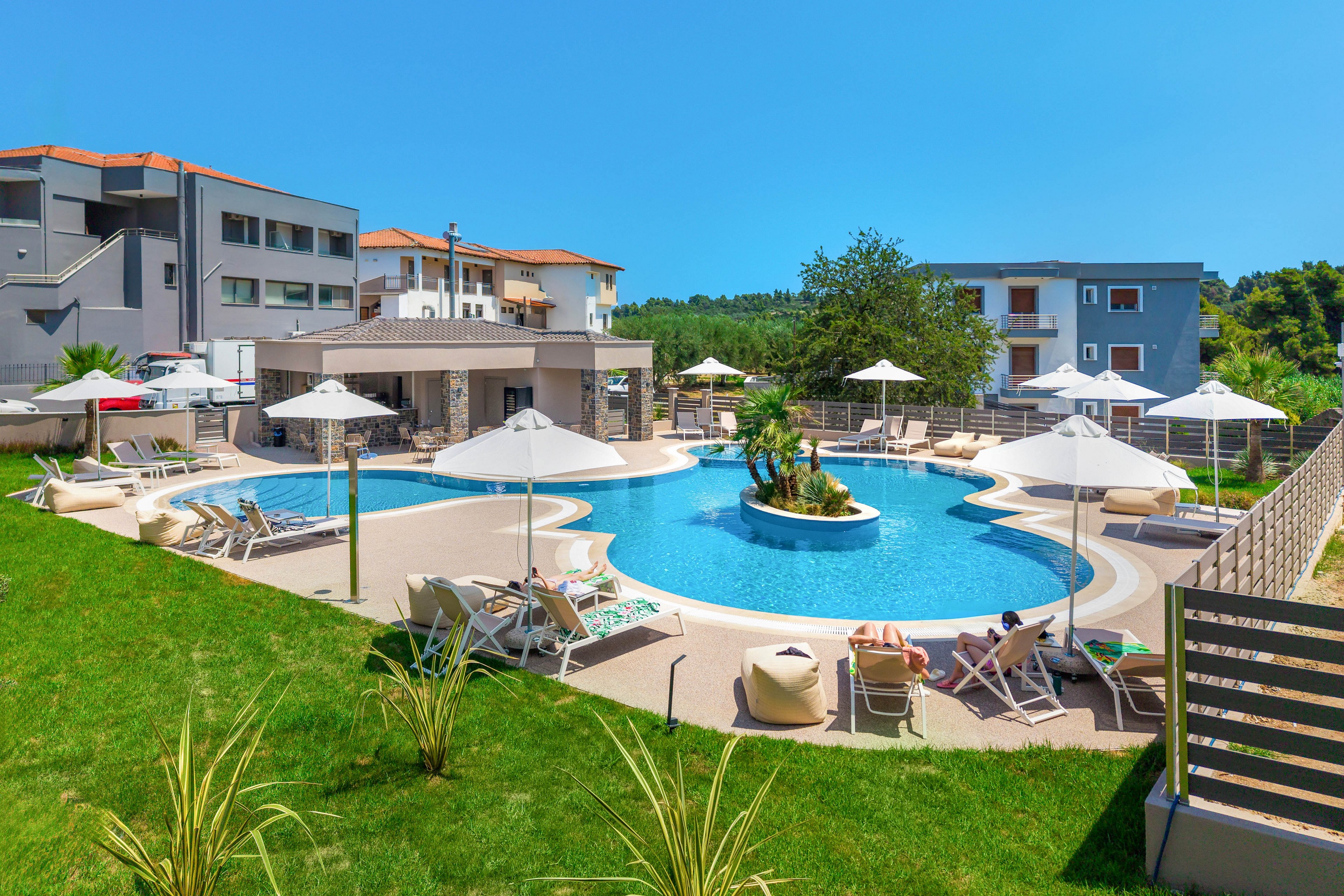 A modern resort features a large circular outdoor swimming pool surrounded by sun loungers and umbrellas. Guests relax on chairs and in the shade, enjoying the bright sunny weather. Contemporary low-rise buildings frame the pool area, with balconies overlooking the scene. Lush green grass and landscaped plants add a tropical feel to the tranquil holiday setting.