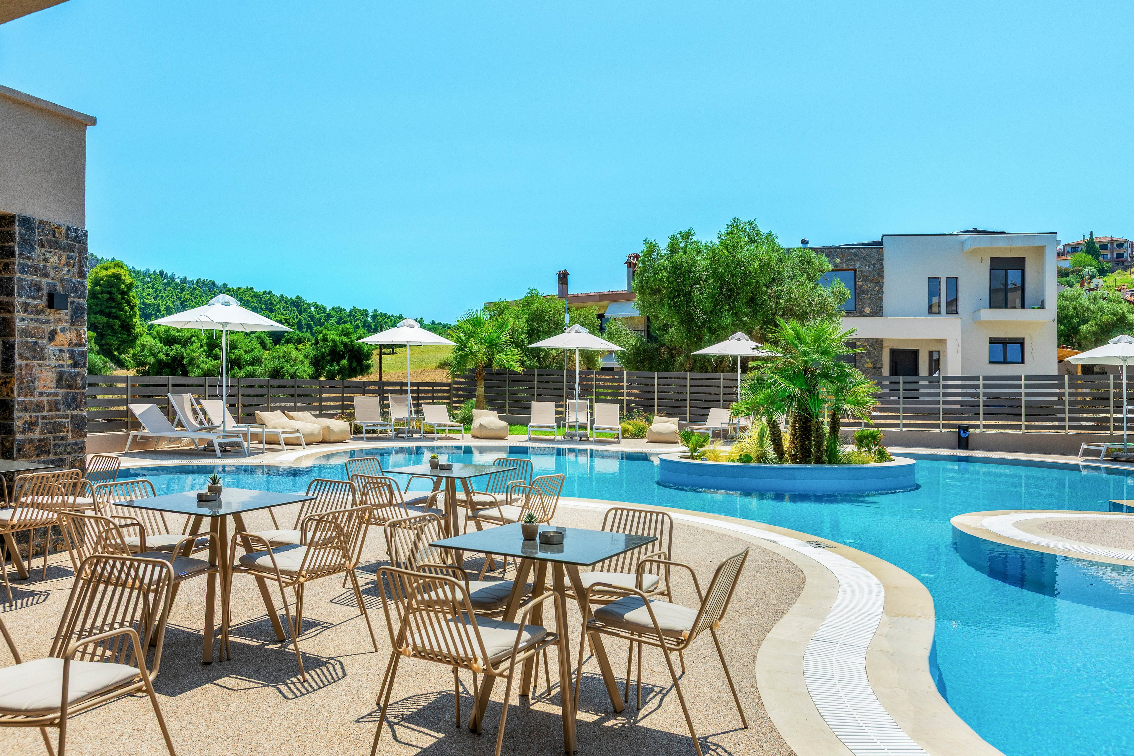 An outdoor resort pool area features metal chairs and tables arranged on a sunlit terrace. A curved swimming pool with clear blue water runs alongside the seating, bordered by a light-colored walkway. White sun loungers and umbrellas line the far side of the pool, with palm trees and greenery adding a tropical touch. Modern low-rise buildings and a bright blue sky complete the relaxed vacation setting.