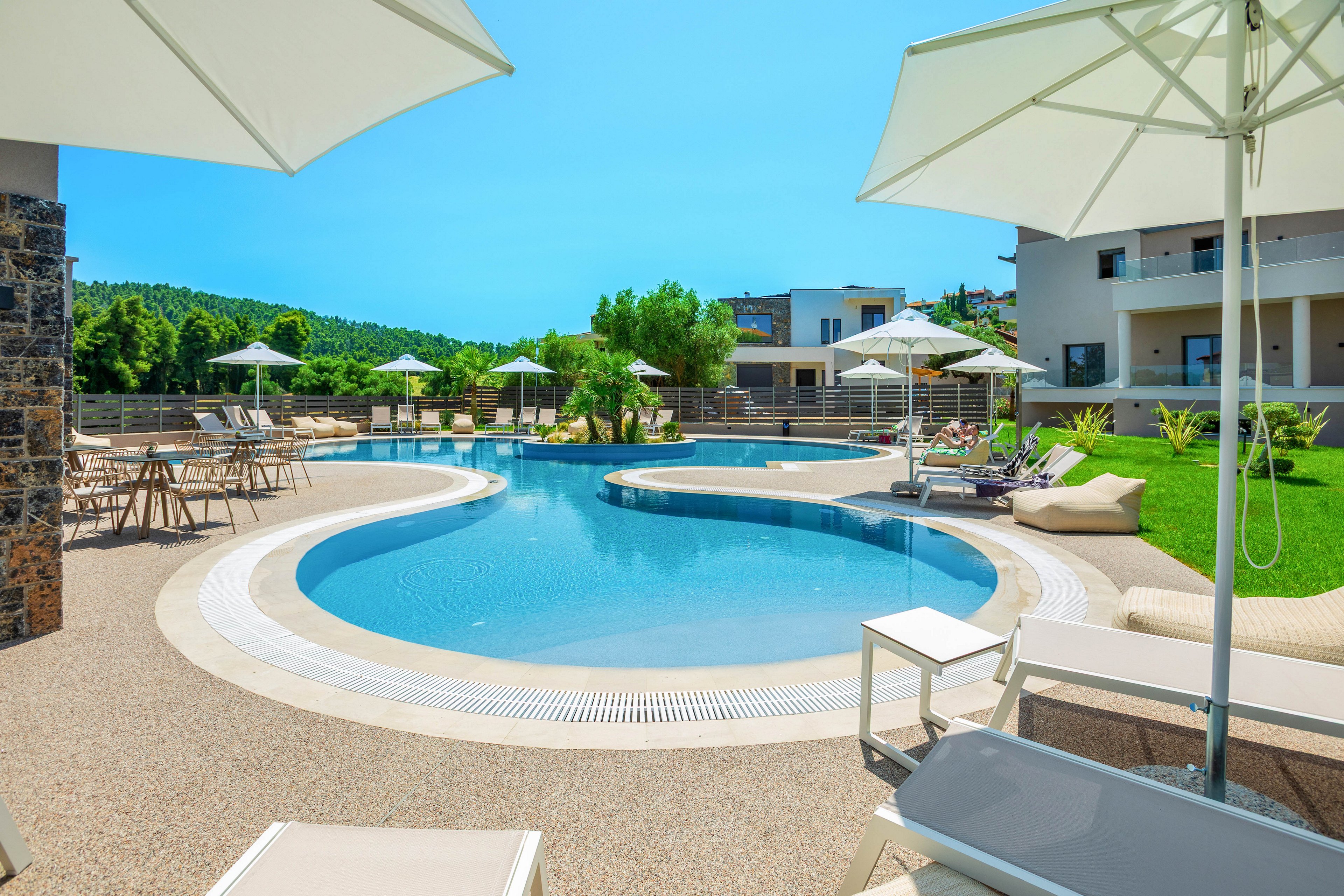 A contemporary resort-style swimming pool curves gracefully through an open courtyard. White sun loungers and umbrellas surround the pool, creating a relaxing leisure area under a bright blue sky. Modern low-rise buildings frame the scene, with green lawns and trees adding natural contrast. The overall atmosphere is calm, sunny, and inviting for vacation guests.