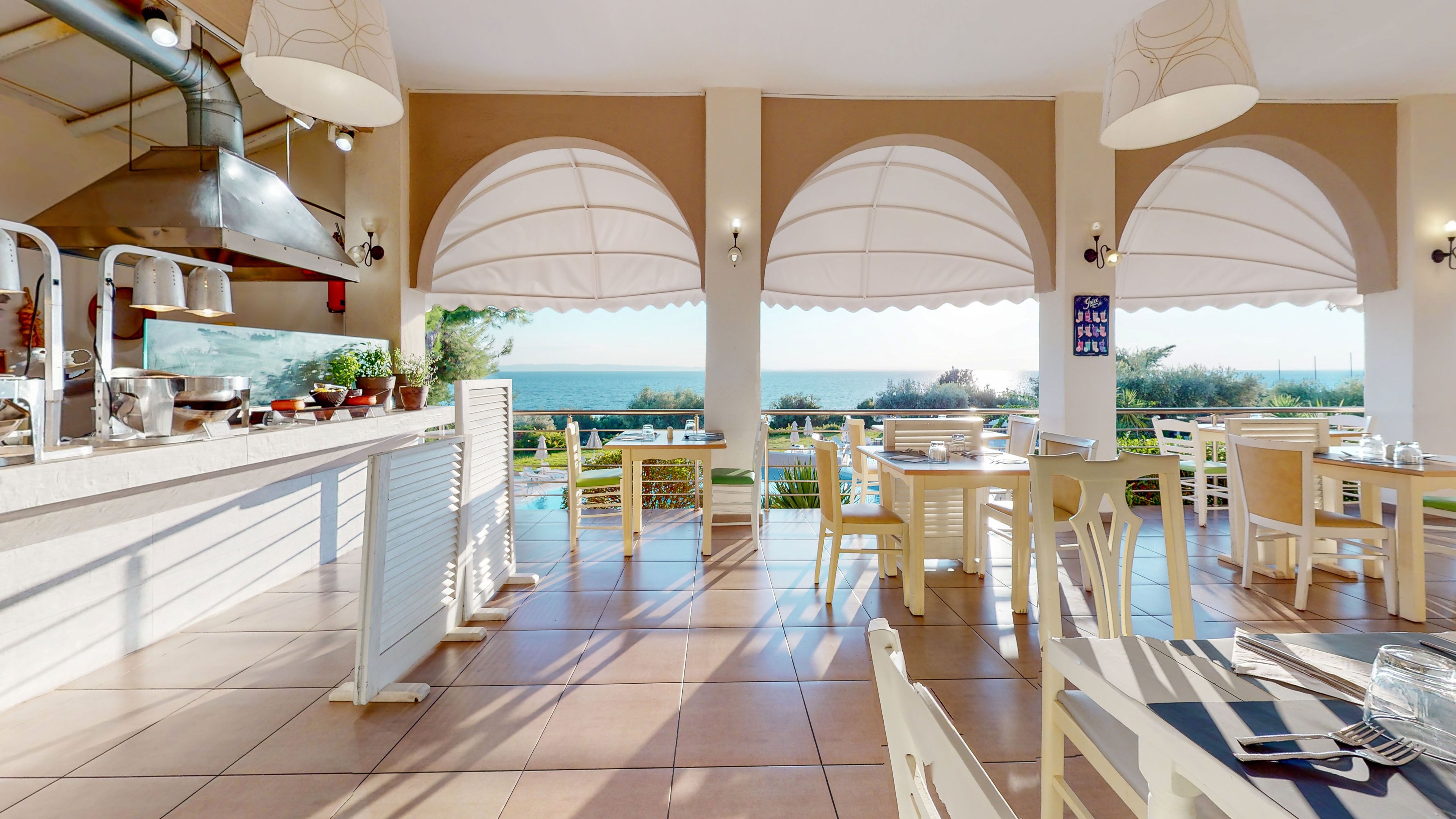 An airy, open-sided restaurant overlooks a scenic ocean and coastal greenery. Light-colored wooden tables and chairs are neatly arranged on a tiled floor, with place settings ready for guests. Large arched openings with white awnings frame the outdoor view, creating a relaxed, vacation atmosphere. A counter with cooking equipment and utensils is visible on the left, suggesting a casual dining or buffet service.