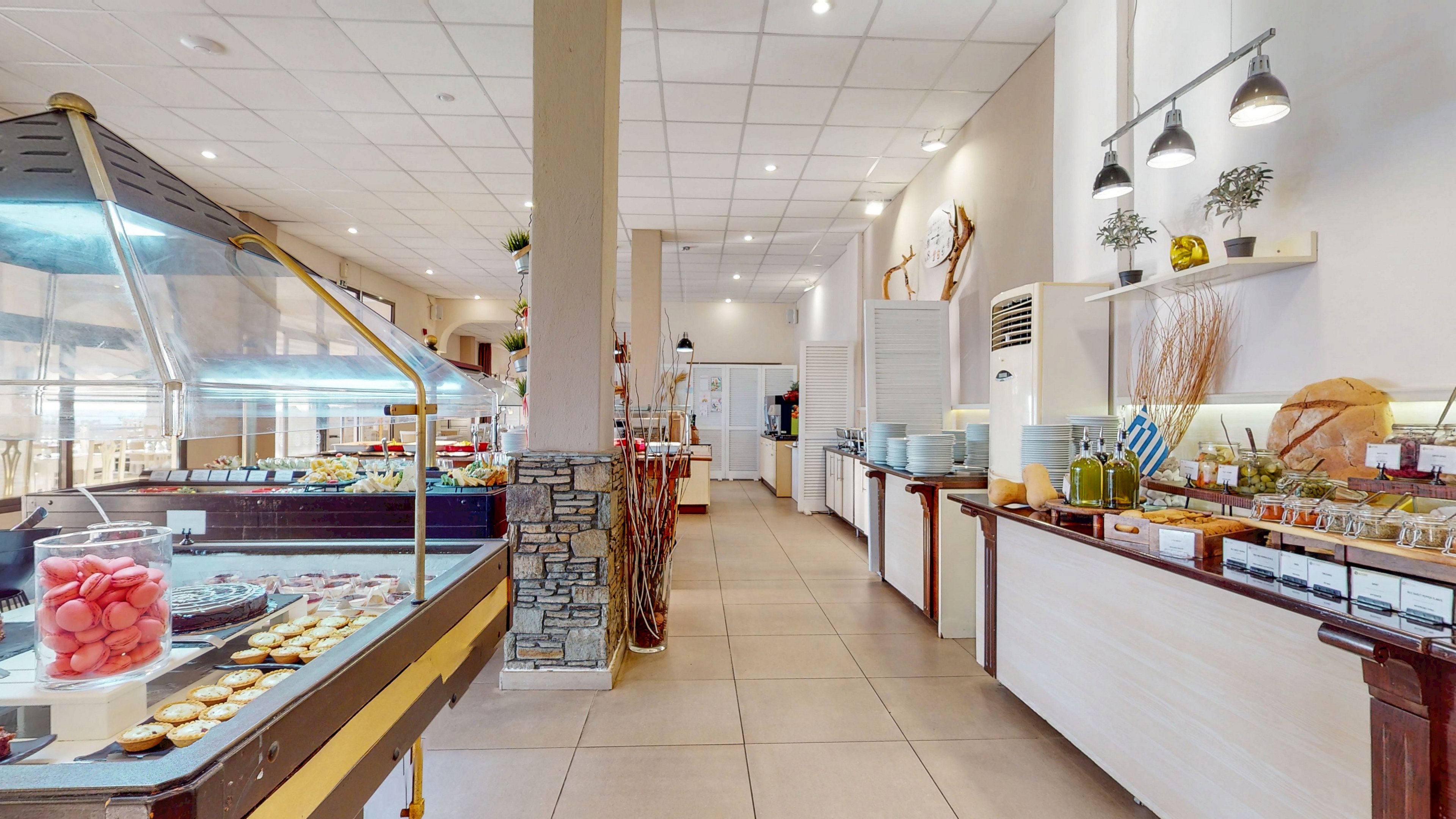 Spacious buffet restaurant interior with multiple food stations and display counters. The setting features a self-service area with desserts, salads, and condiments arranged neatly under bright lighting. Neutral tiled flooring and white walls create a clean, modern atmosphere, complemented by decorative plants and rustic accents. Refrigerated units, beverage dispensers, and serving utensils are visible along the counters, suggesting a hotel or resort dining area.