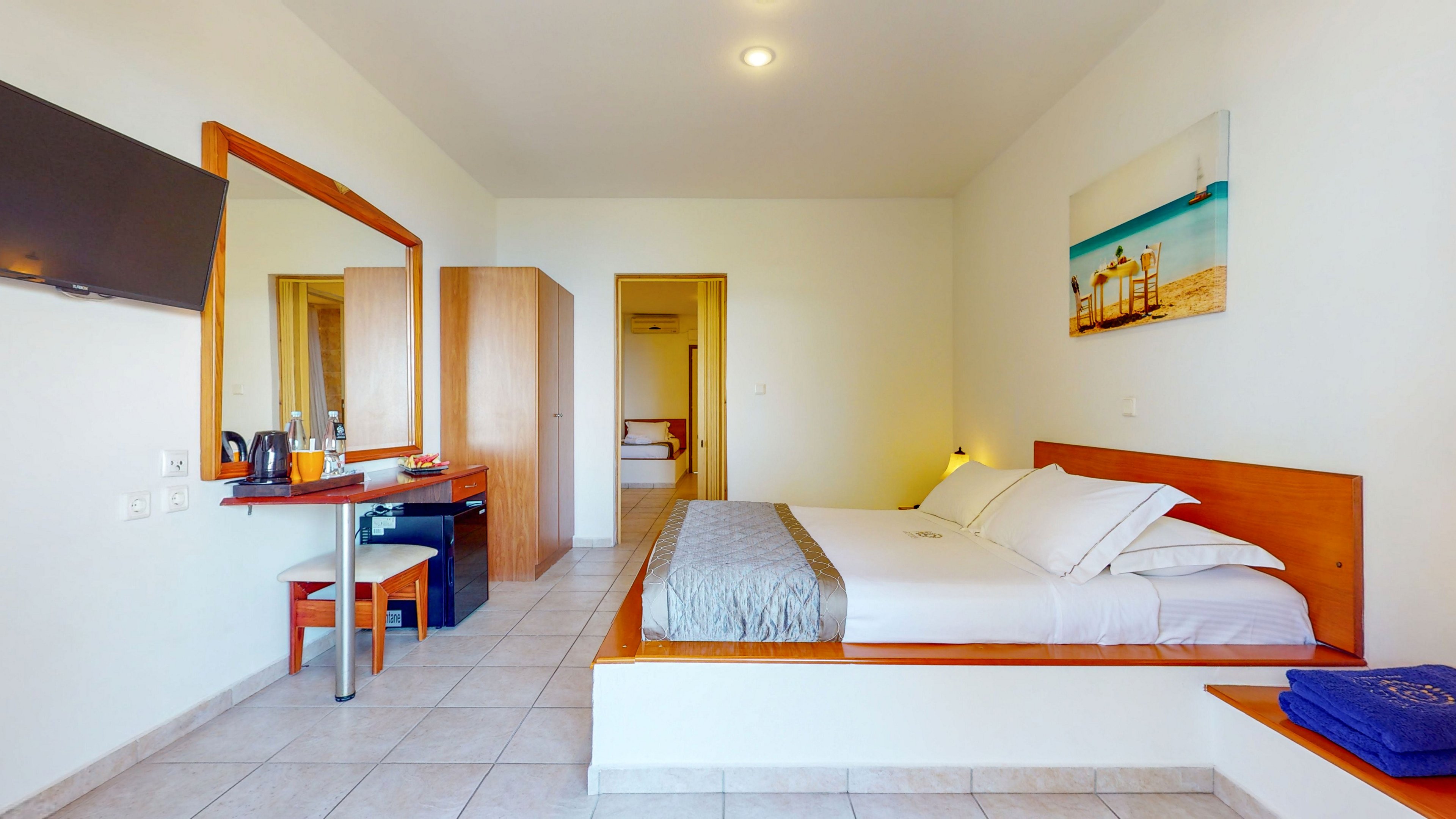 A clean, modern hotel room features a large double bed with white linens and a light gray runner. The space includes a wooden desk with a chair, electric kettle, and mini fridge, as well as a wall-mounted flat-screen television. A wardrobe and an open doorway lead to another area of the accommodation. The room is decorated with a beach-themed wall art and neatly folded blue towels, creating a relaxed vacation atmosphere.