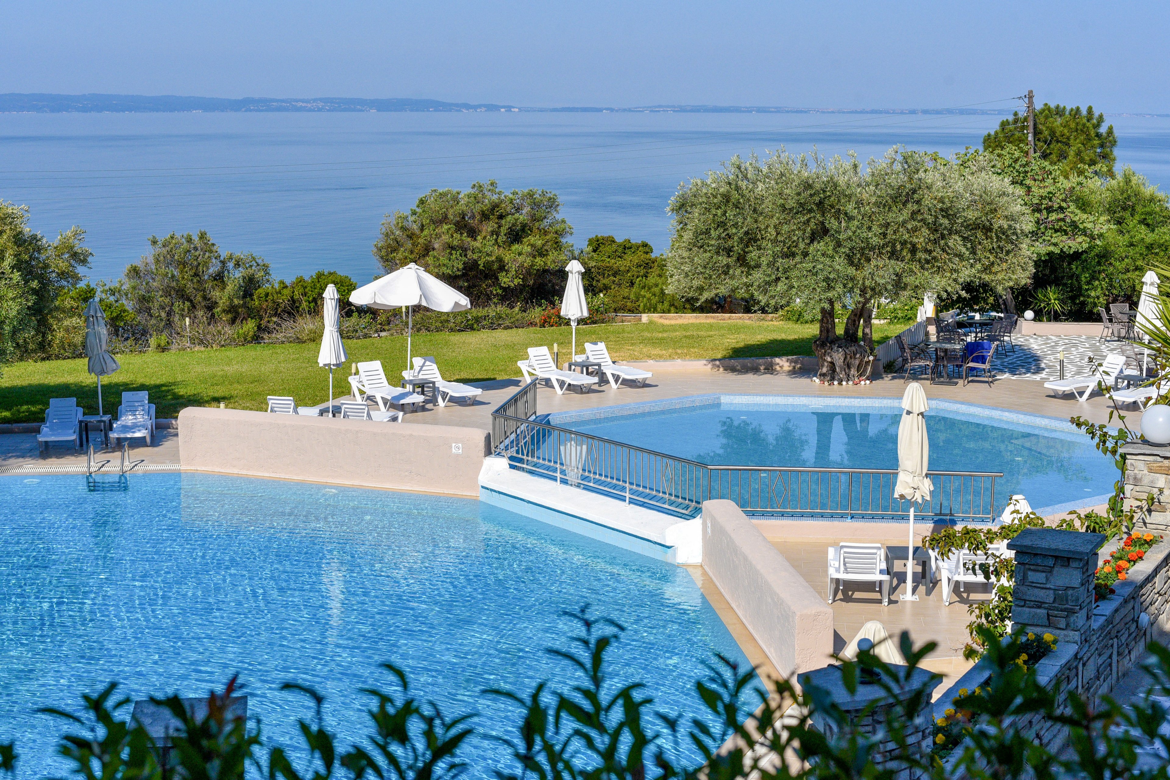 A large, clear blue swimming pool overlooks a calm sea, creating a relaxing vacation setting. White sun loungers and umbrellas are arranged around the pool on a manicured lawn and stone terrace. Green shrubs and mature trees frame the scene, adding natural contrast to the blue water and sky. The overall atmosphere is tranquil and inviting, ideal for leisure and summer holidays.