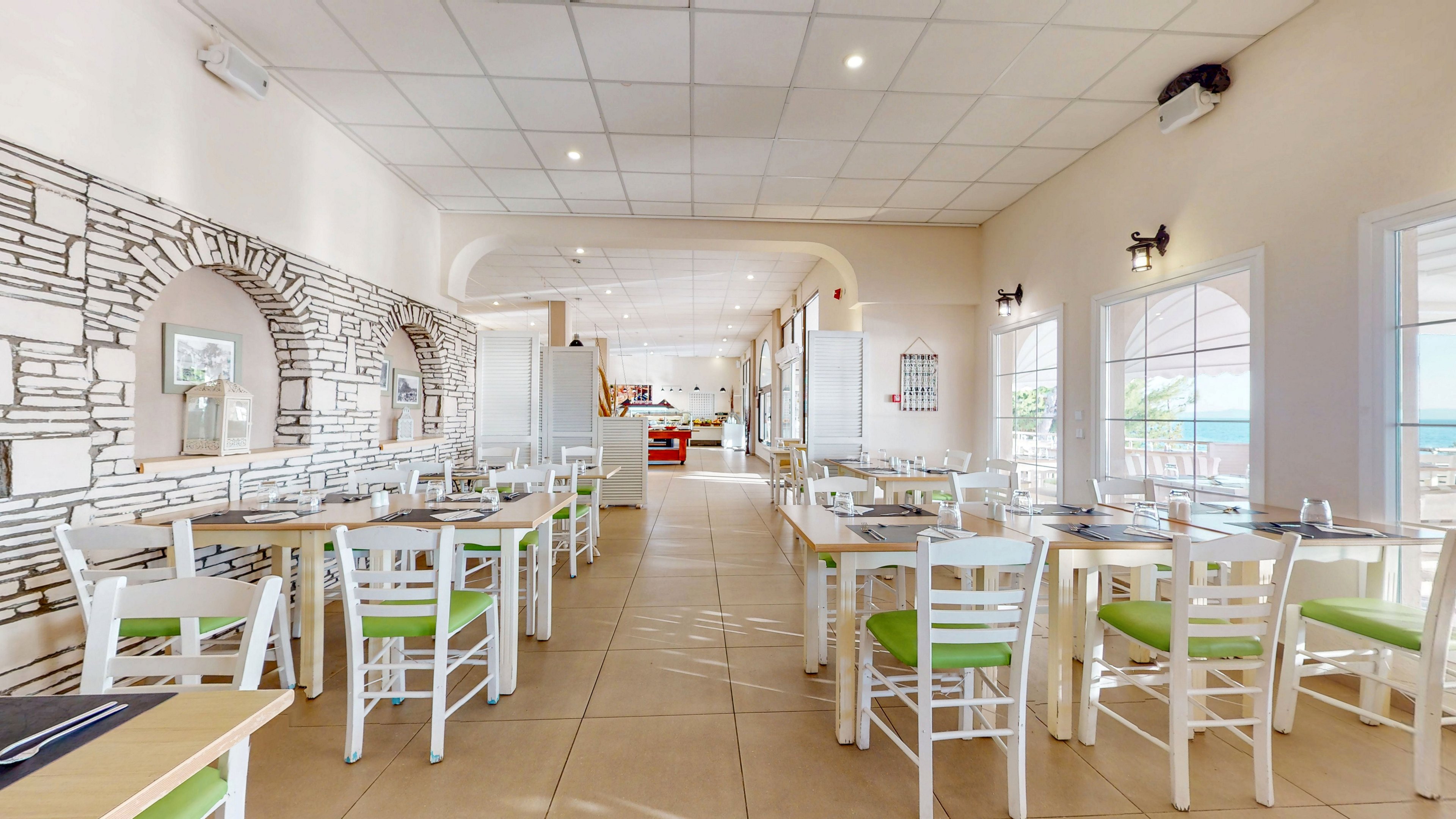 A spacious restaurant interior features rows of white wooden tables and chairs with light green seats. The room is brightly lit with large windows on the right side offering a view of the sea. Stone-pattern wall accents and simple wall lamps add decorative detail to the otherwise minimalist space. The setting appears clean, airy, and ready for guests, with neatly arranged place settings on the tables.