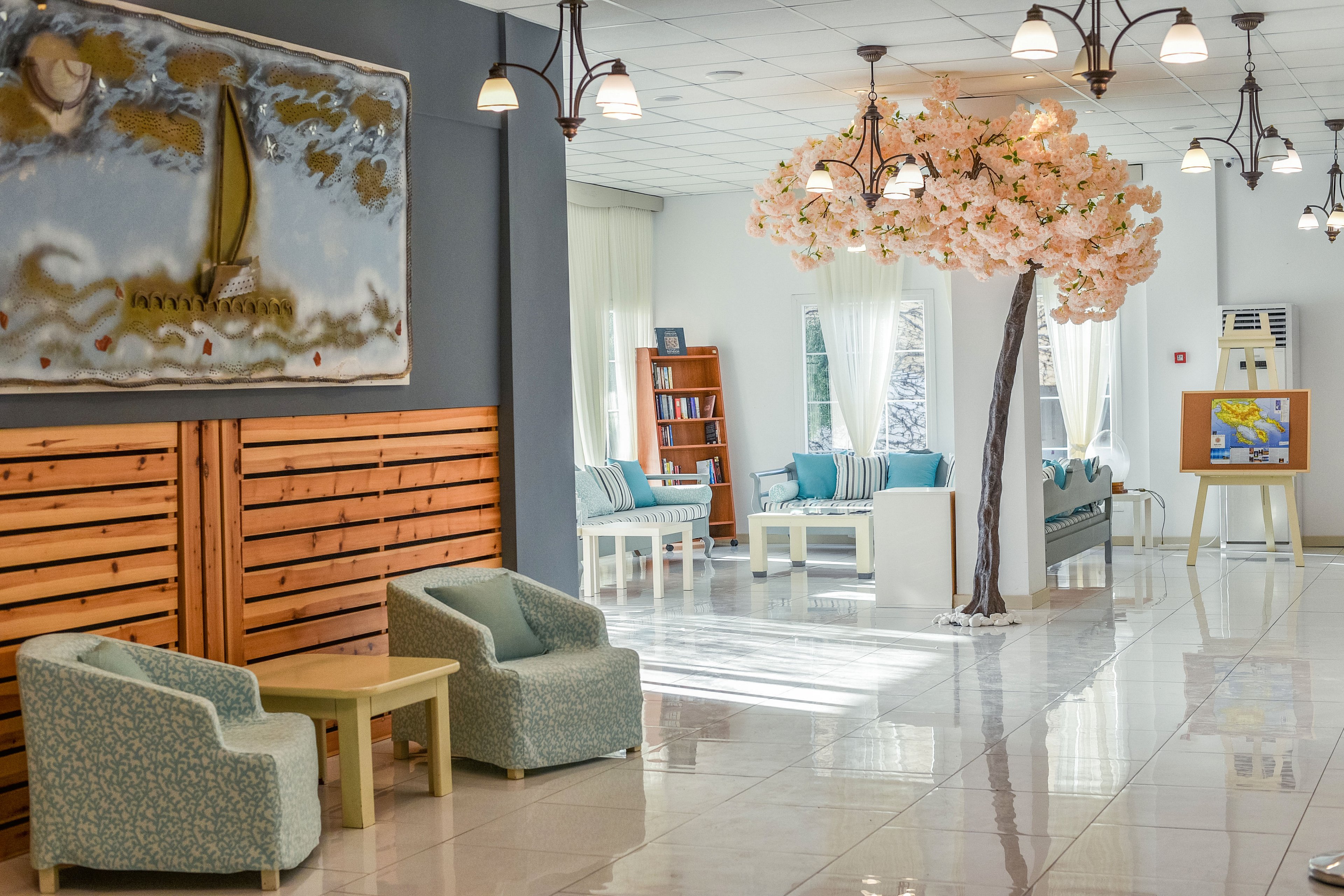 Spacious lounge interior features pastel armchairs and sofas arranged around low tables. A large artificial blossom tree with hanging lights stands in the center of the room. White curtains, tiled floors, and soft blue accents create an airy, coastal atmosphere. Wall art with a sailboat and a bookshelf in the background add visual interest and a relaxed, vacation-like feel.