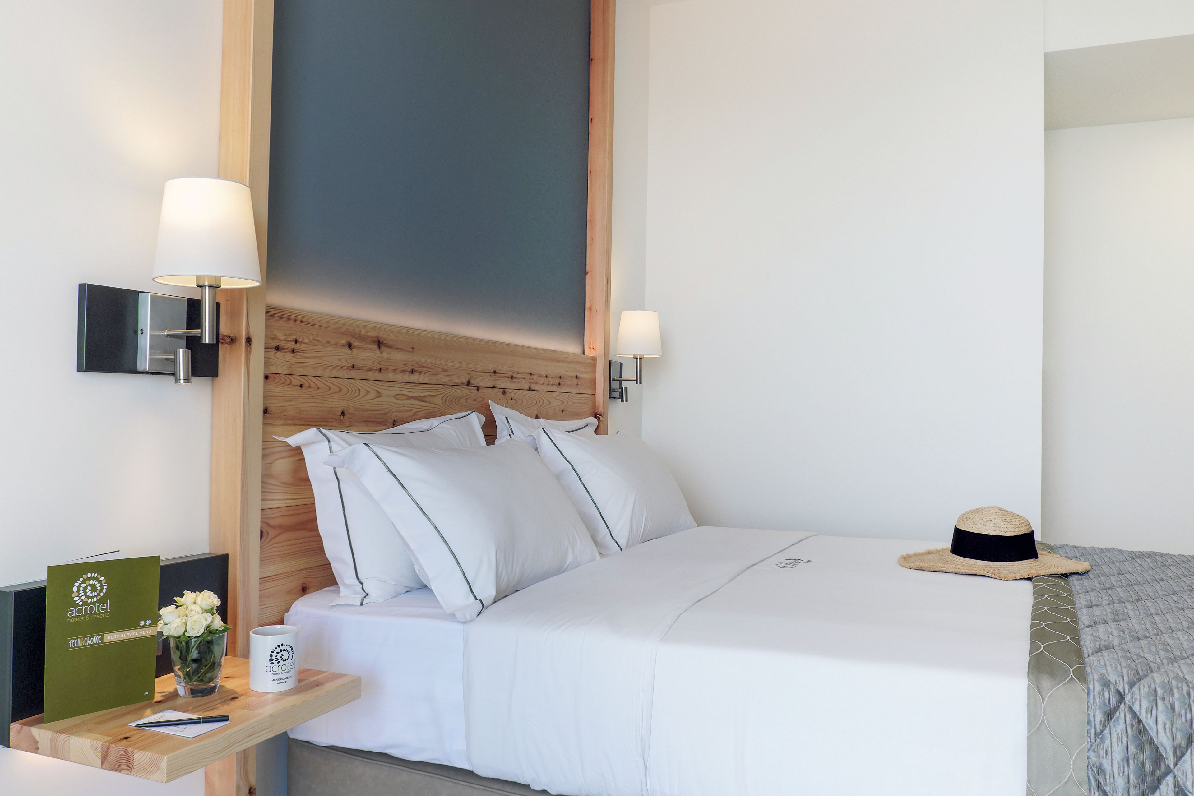 A neatly made double bed with crisp white linens is the focal point of a modern hotel room. Wooden accents, wall-mounted lamps, and a soft gray headboard create a clean, contemporary look. A bedside shelf holds a green booklet, a small framed item, and a vase of white flowers, while a straw hat rests on the bed. The overall atmosphere is calm, inviting, and well-lit, suggesting a comfortable place to relax.