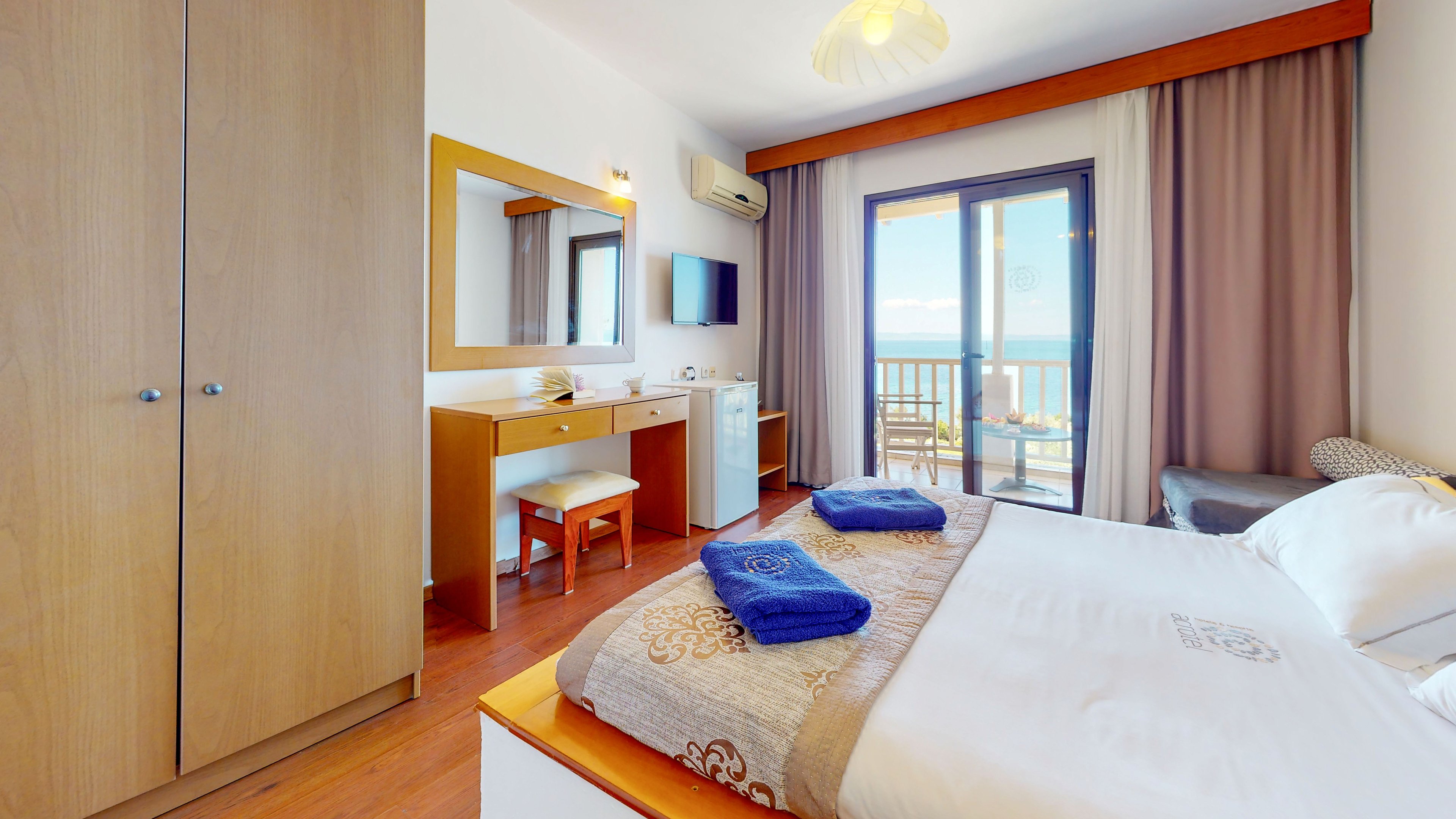 A neatly arranged hotel bedroom features a double bed with folded blue towels on top. The room includes a wooden wardrobe, a dressing table with mirror, a small refrigerator, and a wall-mounted television. Large glass doors open onto a balcony with a view of the sea and coastal landscape. Warm wood tones, white walls, and soft curtains create a clean, inviting atmosphere.