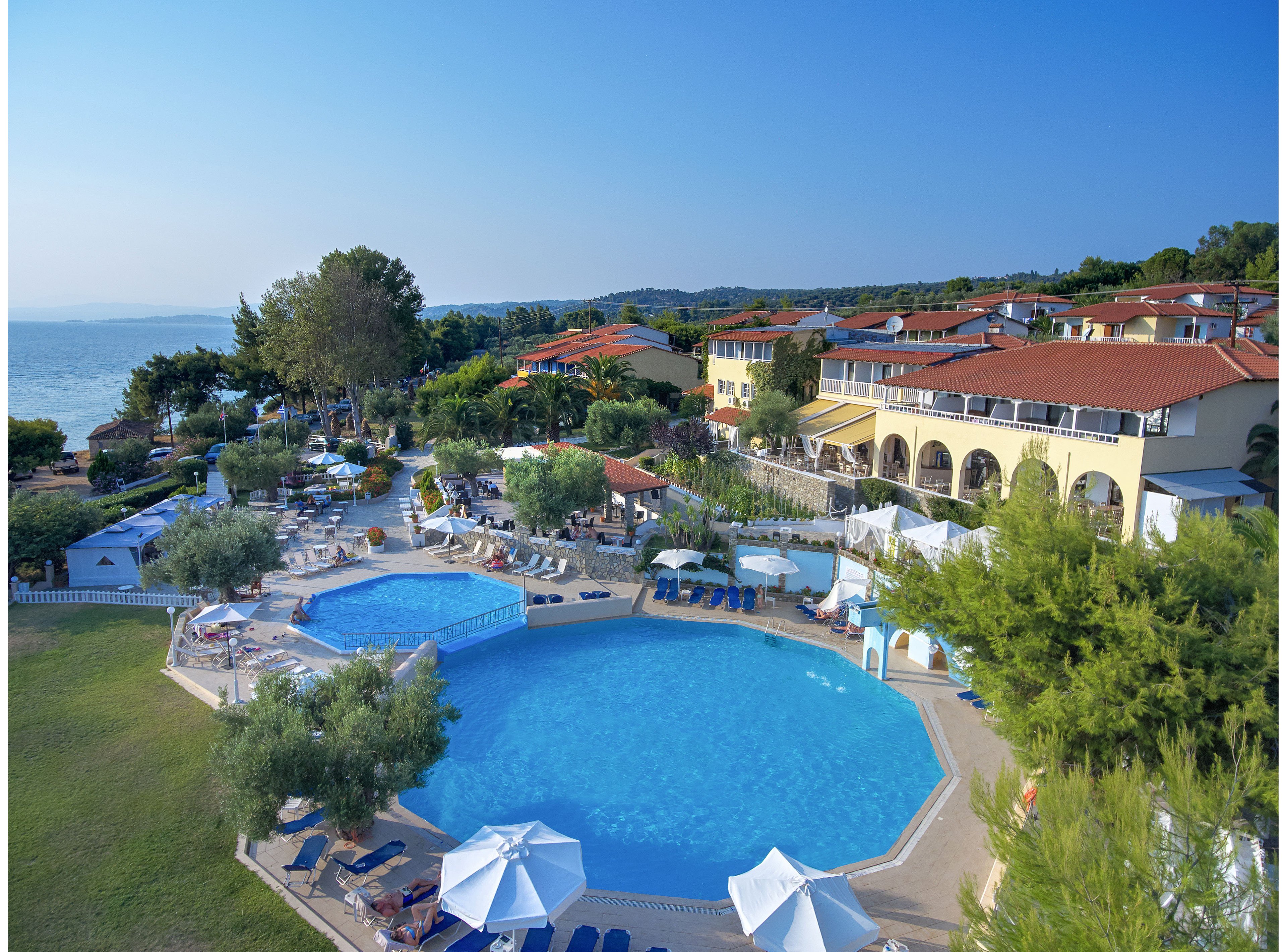 A large coastal resort features multiple outdoor swimming pools surrounded by sun loungers and umbrellas. The complex includes several buildings with red-tiled roofs overlooking the water and landscaped gardens. Guests relax around the pools and seating areas under clear blue skies. The sea is visible in the background, emphasizing the vacation and leisure atmosphere.