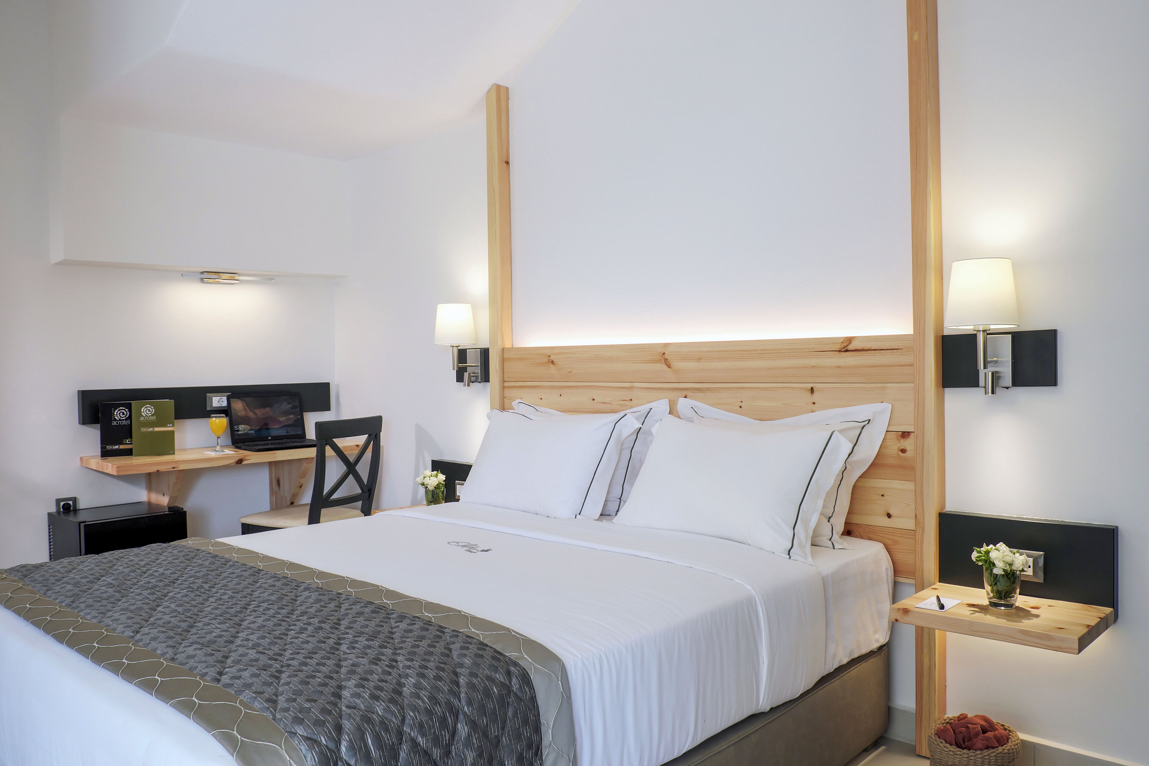 A neatly made double bed with white linens and a light wood headboard is the focal point of this modern hotel room. Soft wall-mounted lamps provide warm lighting on either side of the bed, with small bedside tables holding flowers and decor. In the background, a compact workspace with a chair, desk, and shelving is visible against the white walls. The overall design is clean and contemporary, featuring neutral tones and minimalist furnishings.